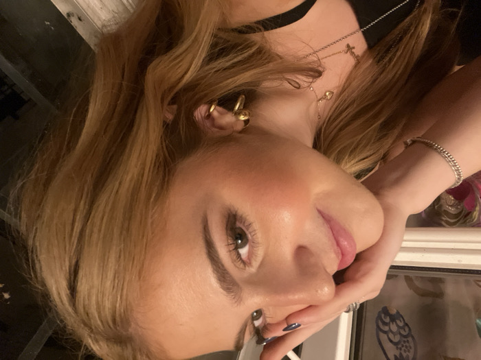 The image features a close-up, angled portrait of a young woman, illuminated by ambient lighting, set indoors. Her head is tilted slightly to the right, and her gaze is directed upwards and to the right of the frame. She possesses fair skin, pronounced blush on her cheeks, and full pink lips. Her eyes are light-colored, appearing green or hazel. Her eyebrows are dark and well-defined. Her long, wavy hair is a reddish-blonde or light brown hue, cascading over her shoulders and partially obscuring the right side of the frame.
Her left hand is positioned against the left side of her face, with fingers resting near her temple and thumb supporting her jawline. Her fingernails are painted with dark polish, appearing dark blue or black. A silver-colored, twisted-chain bracelet adorns her left wrist, and two rings are visible on her left hand's ring and middle fingers. Around her neck, multiple necklaces are visible, including one with a small cross pendant and another with a small heart-shaped pendant. Two gold-colored hoop earrings are worn in her right ear. She is wearing a black garment, likely a top or dress, visible at the bottom of the frame.
The background on the left side shows a blurry reflection or view through a window, revealing indistinct patterns in shades of blue, white, and possibly gold or brown, suggestive of decorative elements or fabric. The background on the right side is dark and lacks discernible details, indicating an interior space. The scene is an indoor setting; however, no specific landmarks or contextual cues are visible to identify a precise location within Paris, France.