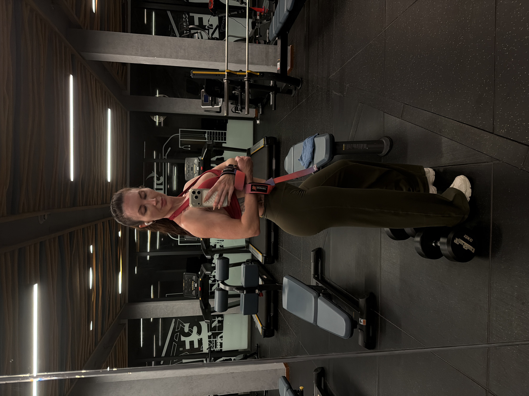 A woman is taking a mirror selfie in a gym. She is wearing a red sports bra and olive green sweatpants. She has her hair pulled back in a ponytail and is wearing white sneakers. She is holding her phone in her right hand and has a pink weightlifting strap on her left wrist. In the background, there are treadmills, weight benches, and weight racks. The gym appears to be modern and well-equipped. The image is taken from the waist up, and the woman's face is partially obscured by her phone. There are no other people visible in the image. The location is not specified in the image or surrounding context.