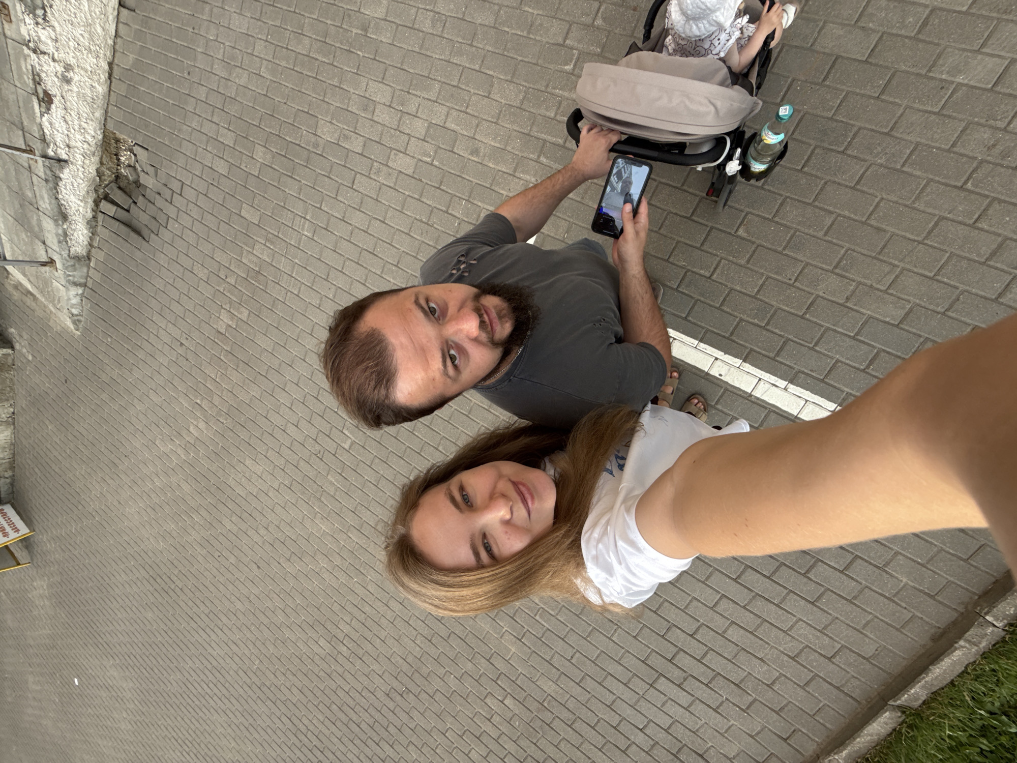 This is a high-angle, full-shot, outdoor selfie taken by a woman holding a phone. She is standing next to a man who is pushing a stroller. The woman has long, blonde hair and is wearing a white t-shirt. The man has short brown hair and a beard and is wearing a dark t-shirt. He is holding a smartphone in his left hand, which is displaying an image. The stroller is light brown and appears to contain a baby wearing a white hat. A green plastic bottle of water is lying on the ground next to the stroller. The background consists of a paved sidewalk with a textured surface, possibly made of interlocking bricks. In the upper left corner of the image, there is a yellow sign with some Cyrillic text visible. The general environment suggests an urban setting. The question regarding the location in the city of Vladimir, Russia cannot be answered from the provided image.