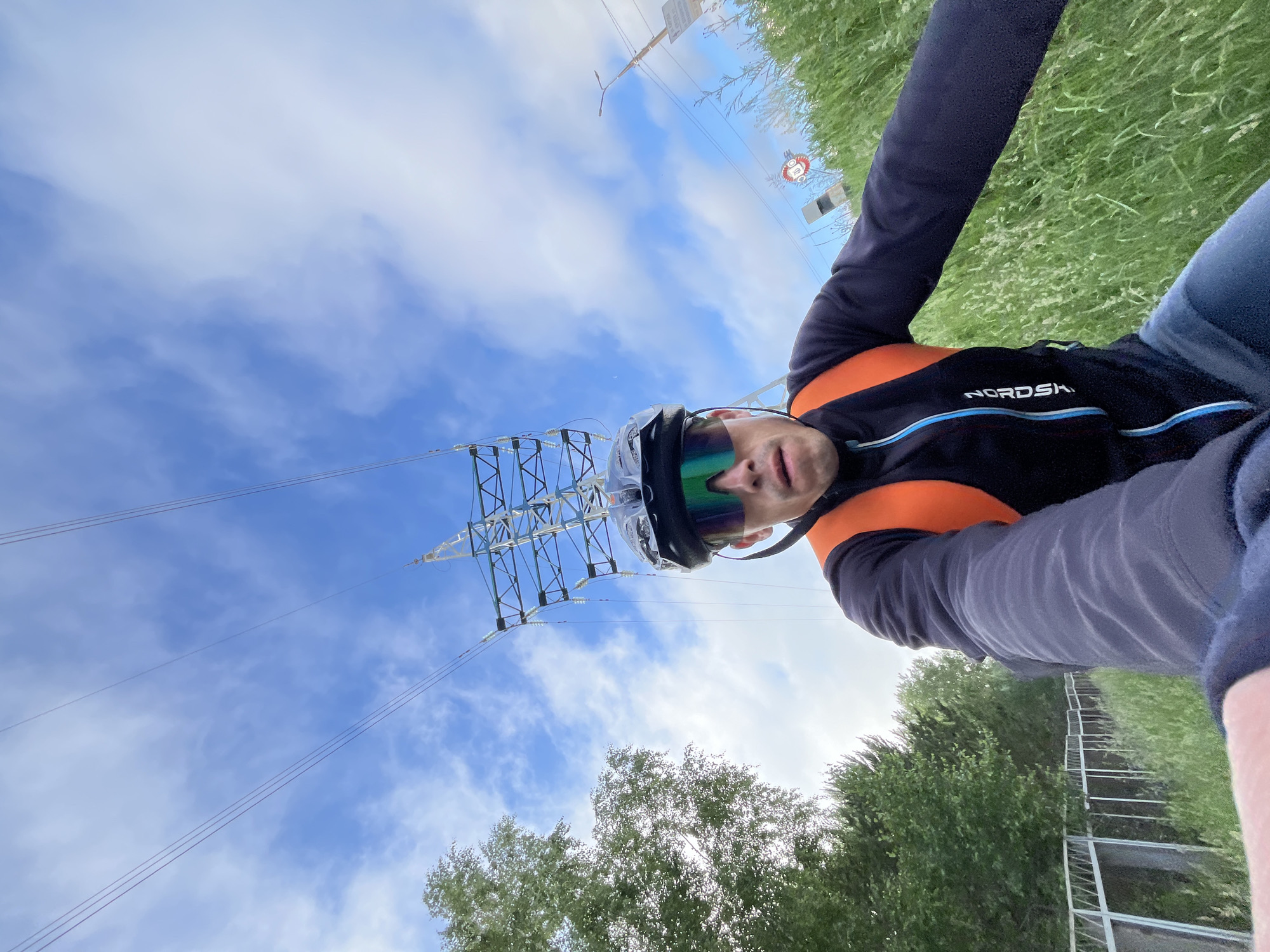 A selfie is taken by a man wearing a black cycling jacket with an orange vest, a helmet, and mirrored sunglasses. He is outdoors, with lush green grass in the foreground and a tall electrical pylon dominating the background. The sky is blue with scattered clouds. To the left, there are trees and a fence. There are also traffic signs visible in the background. The setting appears to be on the outskirts of the city of Chaikovsky, Russia, possibly near a power line corridor.