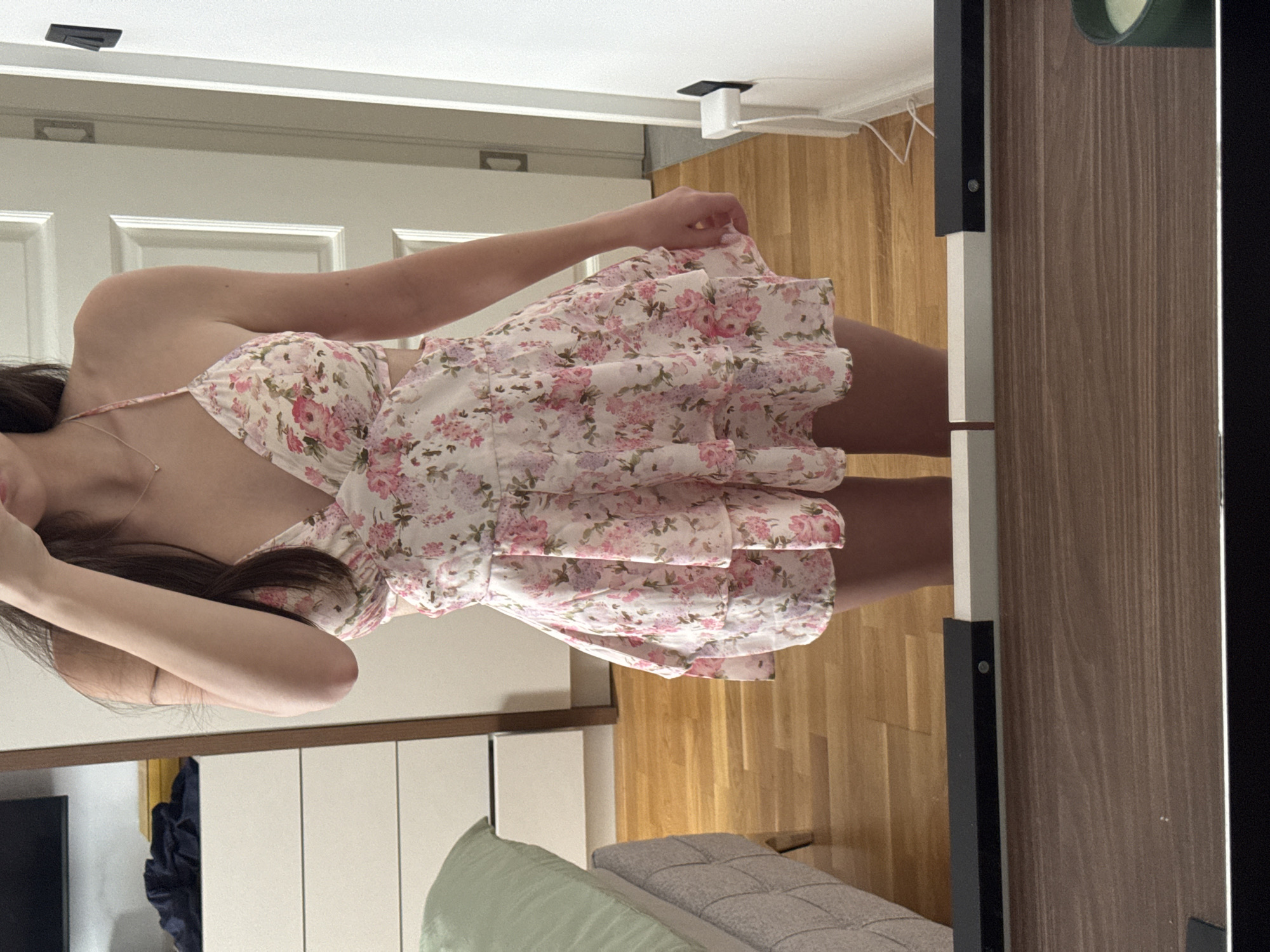 A person is captured in a mirror selfie, or being photographed in front of a mirror, wearing a white, sleeveless mini-dress adorned with a vibrant floral pattern of pink and purple roses and green leaves. The dress features thin straps and a tiered, ruffled skirt. The subject is visible from the chest down, with long dark hair covering part of their face and shoulder, and a delicate necklace around their neck. Their left arm is raised, suggesting they might be holding a camera or posing.
The indoor setting features light wooden floorboards. In the reflection, a bed with a grey quilted frame and a light green pillow is visible. A dark blue item, possibly clothing or a bag, rests near the bed. On the wall behind the subject, a white paneled door is discernible. Above it, a white square device is mounted on the ceiling, with a white cable hanging nearby. The room is brightly lit, indicative of daytime or ample artificial lighting. No text is visible in the image.