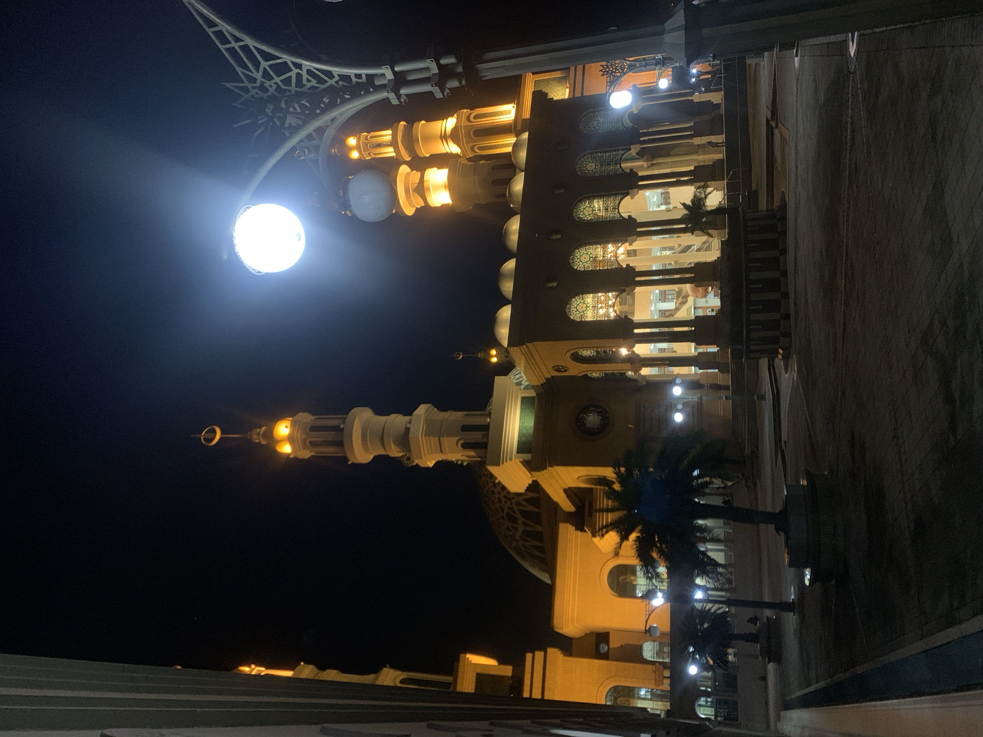 The image presents a grand, illuminated mosque at night, set in Samarinda, Indonesia. The main subject is the mosque's impressive architecture, constructed from light-colored stone or concrete, lavishly lit with warm yellow and white lights that highlight its intricate details.
The mosque features several minarets; one prominent minaret is visible with a golden, crescent moon and star finial. Multiple domes, including a large central dome with elaborate patterns and smaller subsidiary domes, crown the structure. Windows and archways are adorned with decorative screens or patterns, revealing internal illumination and possibly chandeliers.
In the upper left foreground, a geometric, lattice-like structure, likely part of an ornate lamppost or architectural feature, is partially visible. A very bright, circular white light source, appearing to be a powerful floodlight or streetlamp, emanates intense light in this area. Palm trees are seen in the midground and foreground, their dark silhouettes contrasting against the brightly lit mosque and the dark night sky. The ground consists of paved areas, possibly courtyards or pathways.
The scene captures a clear night with no discernible weather events. No people are visible, nor is any specific activity taking place beyond the stationary presentation of the building. The image conveys a sense of peaceful reverence and architectural splendor. No text is visible in the image.