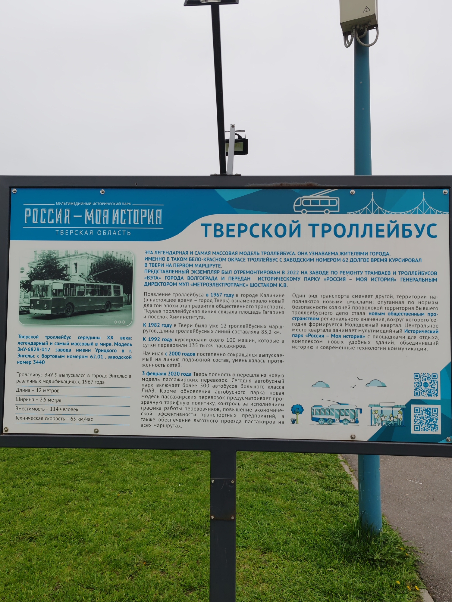 The image shows an information board located in Tver, Russia, specifically dedicated to the history of the Tver trolleybus system. The board is predominantly blue and white, featuring a photograph of a vintage trolleybus, text in Russian, and QR codes. The text provides historical context, including the trolleybus's introduction in 1967, its peak in 1982 with 12 routes, and its eventual replacement by buses in 2020. The sign also mentions the renovation of a historic trolleybus at a factory in Volgograd.