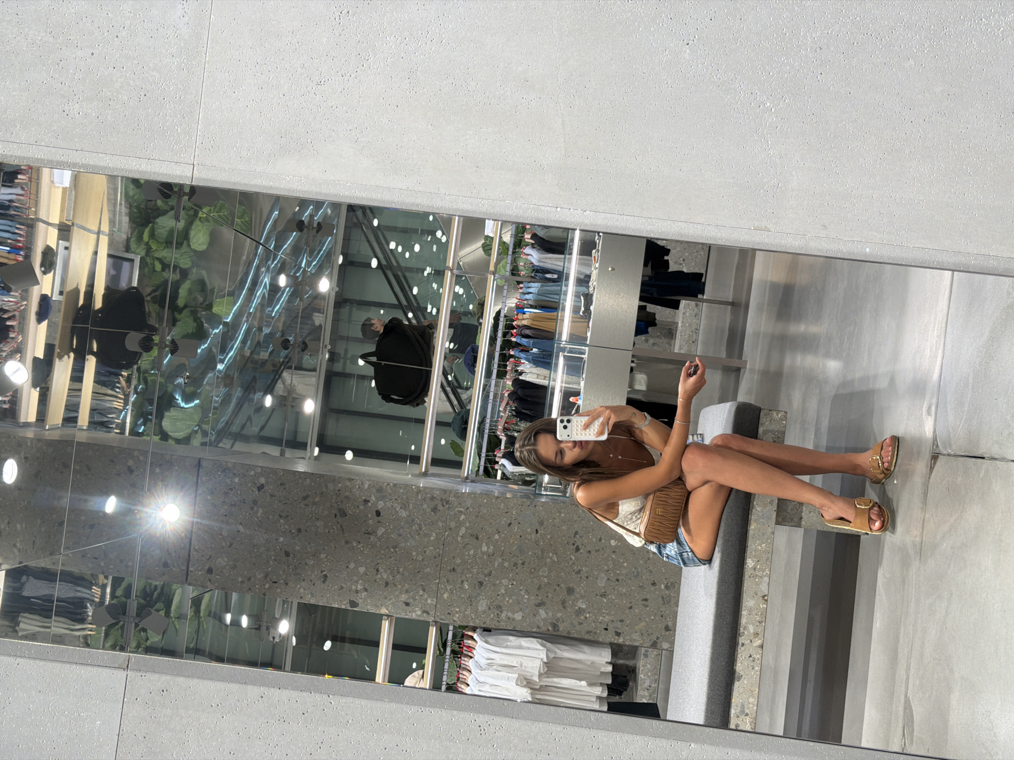 This is a full-body, mirrored selfie of a young woman in a clothing store. The woman is sitting down, holding her phone up to take the photo. She is wearing a white lace tank top, denim shorts, and tan slide sandals. She has a small brown crossbody bag slung over her shoulder. Her expression is relaxed and she appears to be casually browsing.
The setting is a modern retail environment, likely a boutique or a department store, given the racks of clothing and display shelves visible in the reflection. The store features a polished concrete floor and walls, with large mirrored surfaces that create a sense of depth and space. The lighting is bright and artificial, suggesting an indoor setting, possibly during the day or evening.
In the background reflections, other shoppers and merchandise are visible, including clothing on hangers and neatly folded stacks of garments. There are also architectural elements such as escalators and light fixtures. The overall impression is one of a trendy and well-lit shopping space.
There is no visible text in the image. The time of day and weather are not discernible. The location cues suggest an urban retail environment, consistent with the provided context of Milan, Italy.