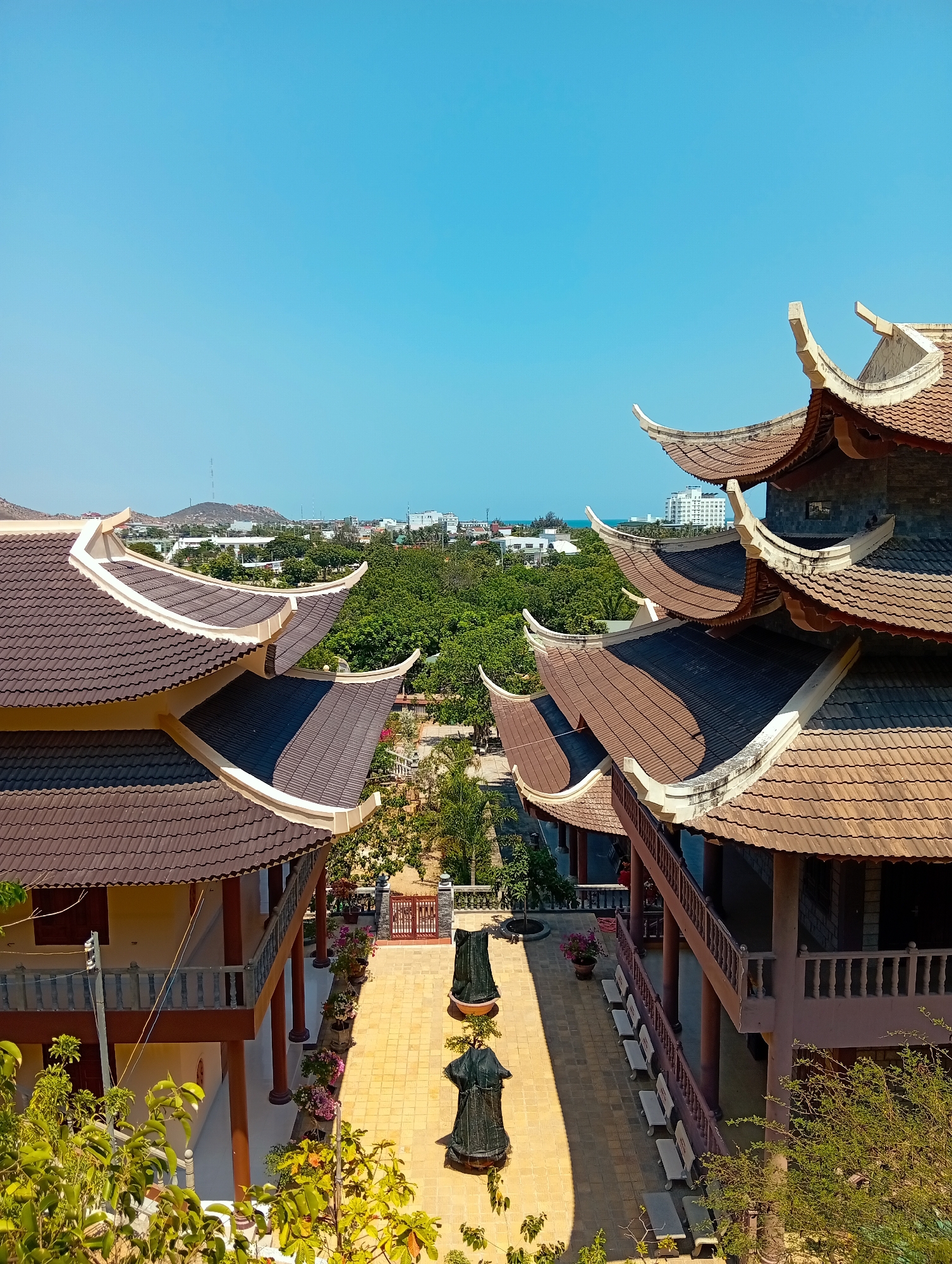 The image shows an elevated view of a courtyard between two buildings, presumably within a temple complex. The buildings feature traditional East Asian architectural elements, including multi-tiered roofs with upturned eaves. The roofs are brown tile with light-colored trim. Balconies with railings are visible on both structures.
In the courtyard, a brick-paved walkway runs between the buildings. Several dark statues stand along the walkway. There are flower pots with flowering plants on either side of the walkway, as well as benches, and a decorative gate is present near the back of the courtyard.
Beyond the temple complex, a cityscape is visible, with various buildings and trees. A body of water appears on the horizon. A bright blue sky is overhead.
Given the location, the complex is likely near the city of Khánh Hải, Vietnam.