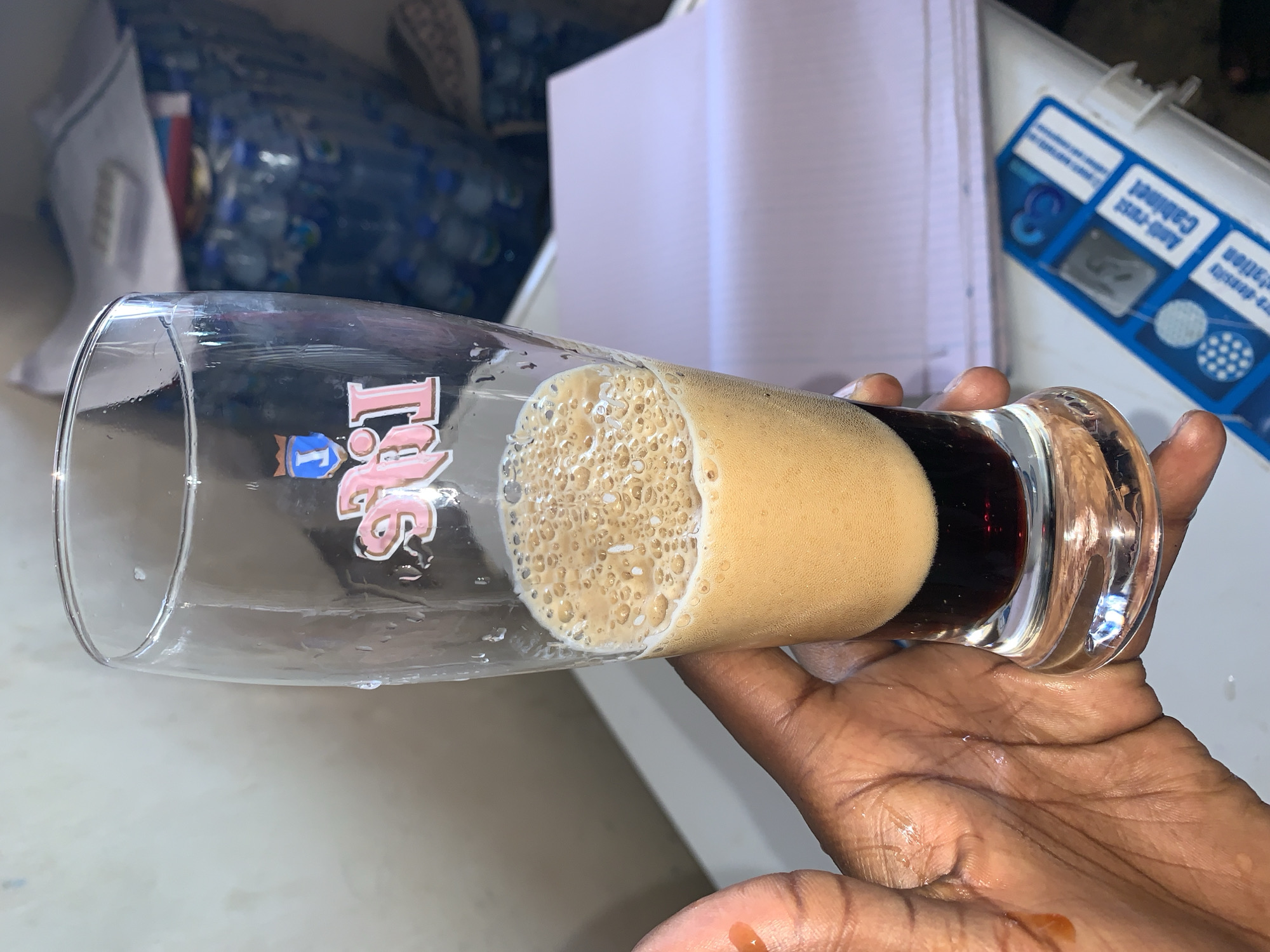A dark-skinned hand holds a tall, clear glass horizontally, filled with a dark brown beverage and topped by a thick, light brown foam. The glass features a distinct logo on its side, displaying "Ting" in stylized pink letters alongside a blue and white emblem. Small droplets of the dark liquid are visible on the hand's palm, suggesting recent handling or pouring.
The background is blurred, indicating an indoor setting. Visible items include a stack of clear plastic water bottles and a white bag or packaging in the upper left. Towards the upper right, an open, light-colored notebook with ruled pages is discernible. Further right, a blue and white object, possibly a component of an appliance or a water filter, shows partially readable text, including "Anti-rust Cabinet," "filtration," and the number "3." The scene portrays a casual moment in what appears to be a domestic or informal environment. While the location context is New Stadium, Nigeria, no explicit visual cues within the image confirm this. No specific time of day or emotional expressions are evident.