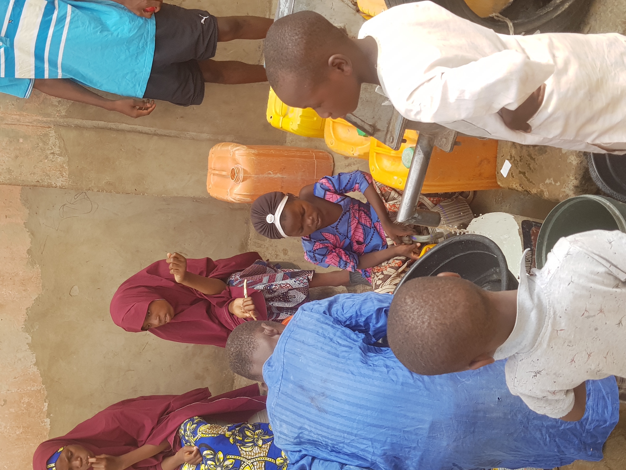 In Dutsinma B, Nigeria, a group of children gathers around what appears to be a water source. Several yellow and orange jerrycans are scattered on the ground. A child wearing a blue and pink patterned dress with a white headband is operating a pump or faucet. Other children watch. Two girls in burgundy hijabs are present. One boy is wearing a blue striped shirt with black shorts. Another child wears a white shirt. A large black bucket is visible.