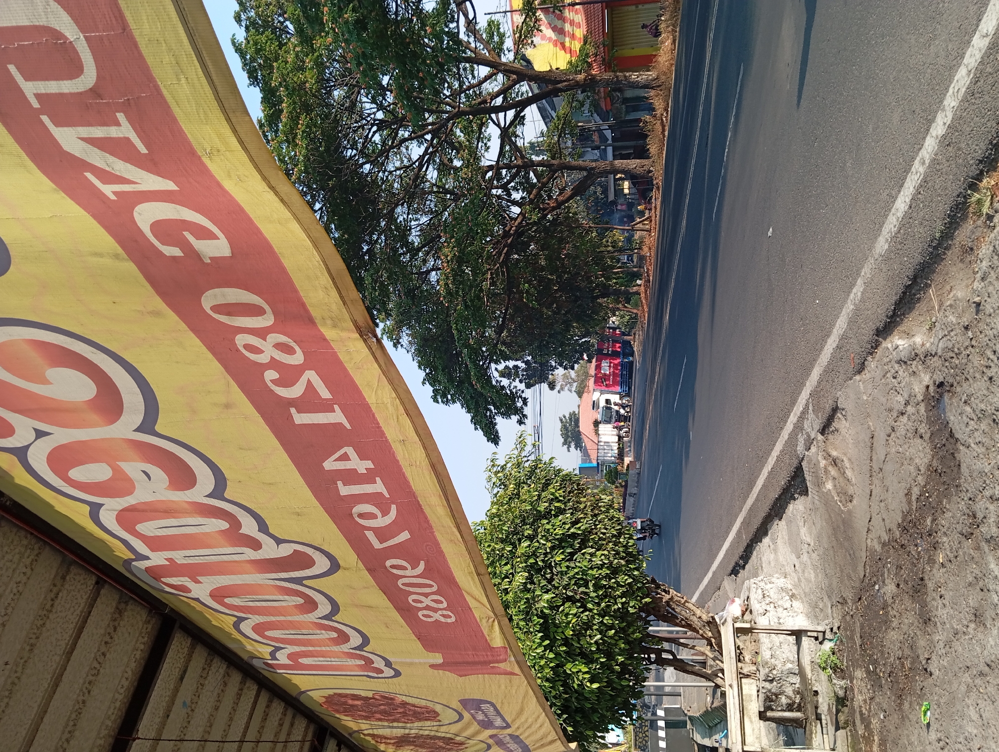 The image captures a street scene in Pandaan, Indonesia, dominated by a large yellow banner with red lettering stretched across the foreground. The banner appears to be a promotional advertisement, with the visible text featuring large, stylized numbers and letters that are partially obscured or angled. The lettering is in red with a white outline.
Behind the banner, a paved road runs horizontally, with a distinct white line marking its edge. The road is relatively clear of traffic, with a few motorcycles visible in the distance. On the left side of the road, there is a sidewalk area with concrete and a small tree. A stone structure is also present on the sidewalk. Further down the road, on the right, are buildings with colorful facades, typical of commercial establishments. Several trees, with lush green foliage, line the roadside, casting shadows on the street. The sky is clear and blue, indicating a bright, sunny day. There are also a couple of trucks parked or driving on the road in the mid-ground. One person is seated on the right side of the road near one of the buildings. The overall impression is a daytime scene on a commercial street in a town or city.