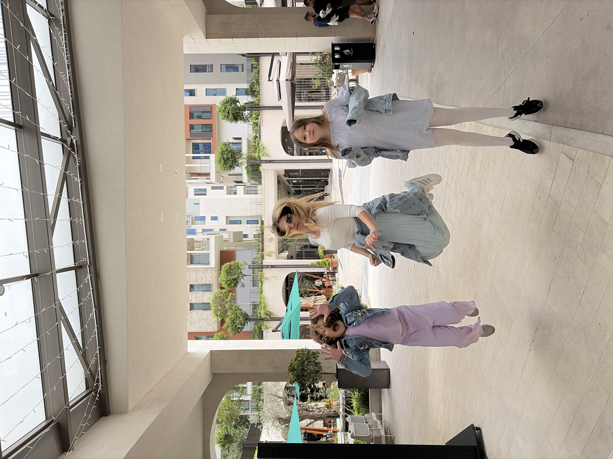 Three individuals are seen on a light-colored tiled floor in what appears to be a covered walkway or outdoor mall. The backdrop reveals buildings of varied heights and architectural styles, indicative of urban planning. The environment is sunlit. The photograph appears to have been taken in Porto Montenegro, Tivat, Montenegro.
The closest individual, facing the camera, is a young person with their hands positioned playfully near their face. They are wearing a lilac-colored outfit and a denim jacket. In the middle of the frame, another person is turned partially away, their face visible, adorned with sunglasses, and clad in a white top and light blue bottoms. The farthest individual is a young girl wearing a striped dress with white stockings and black shoes. The two other people are likely adults. The subjects may be interacting, though the exact nature is difficult to determine from the image.