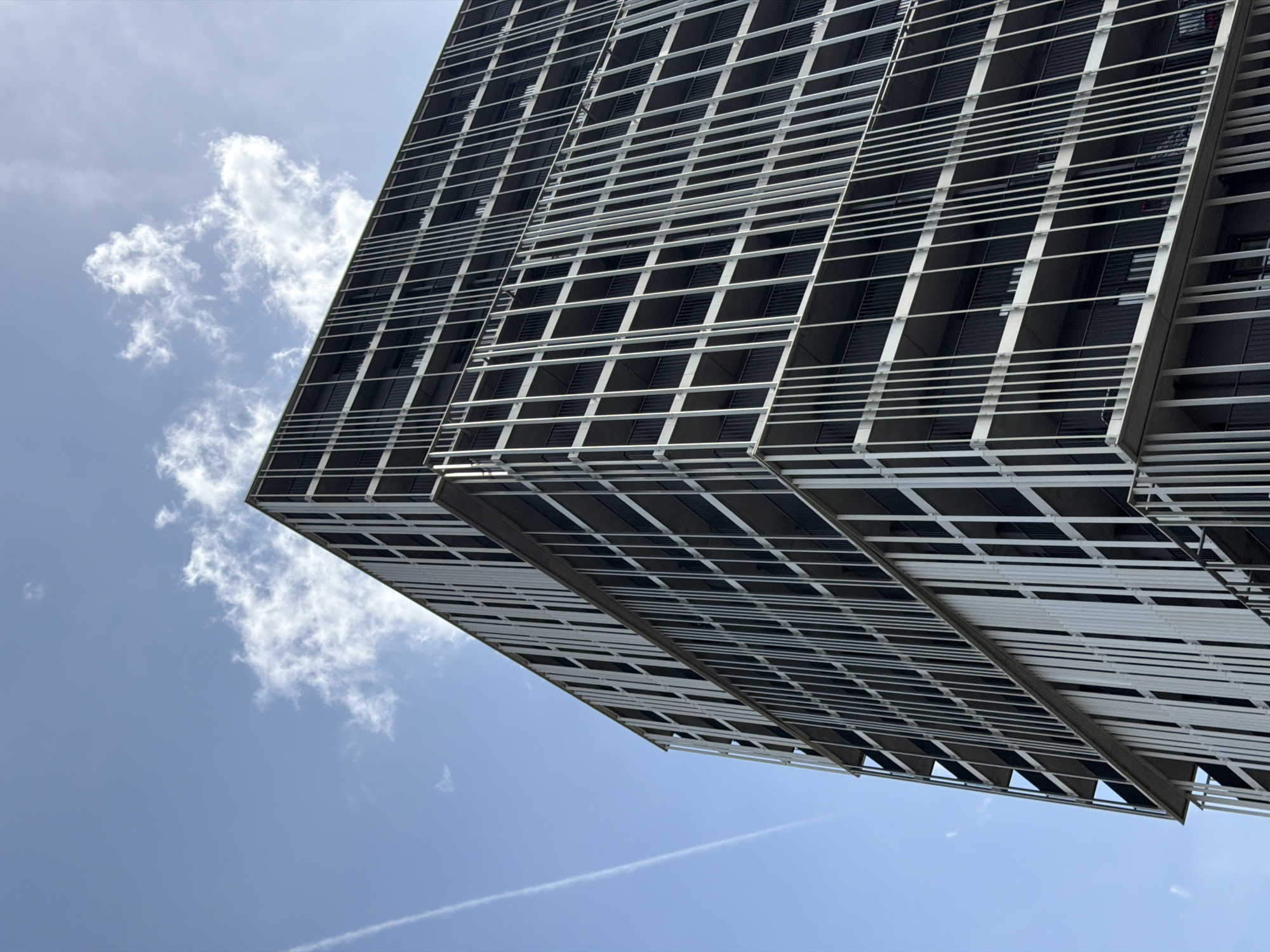A low-angle shot of a building in Esch-sur-Alzette, Luxembourg, features a distinctive facade consisting of a grid-like pattern of white horizontal and vertical lines forming rectangular frames. The frames appear to be filled with dark or shadowed elements, giving the building a textured look. The building's corner is visible, and the structure appears to be multi-storied. To the left of the building, there are scattered white clouds against a blue sky. A contrail is visible in the lower part of the sky. The sky is clear and light blue.