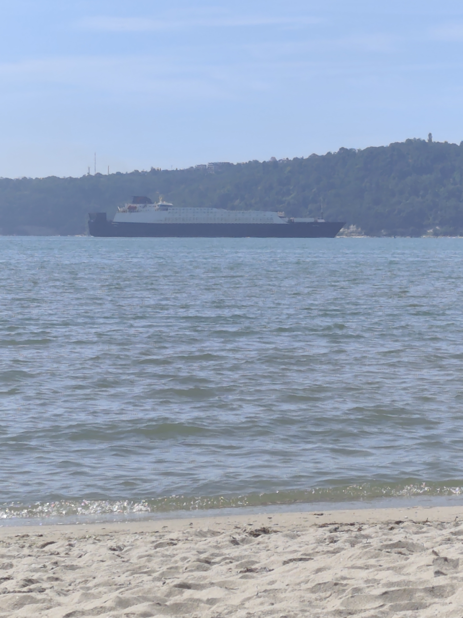 A large cargo ship, possibly a ferry, is visible on the sea, moving from left to right. The ship is dark in color with a white superstructure and appears to have multiple decks. In the background, a densely forested hill rises from the coastline. Several structures, including what look like radio towers and buildings, are present on the hill, suggesting an urban or suburban area. The foreground shows a sandy beach with gentle waves lapping at the shore. The sky is clear and blue with a hazy horizon. This scene is likely captured along the coast of Varna, Bulgaria, known for its beaches and proximity to hills and maritime activity. There are no people or immediate actions depicted in the image.