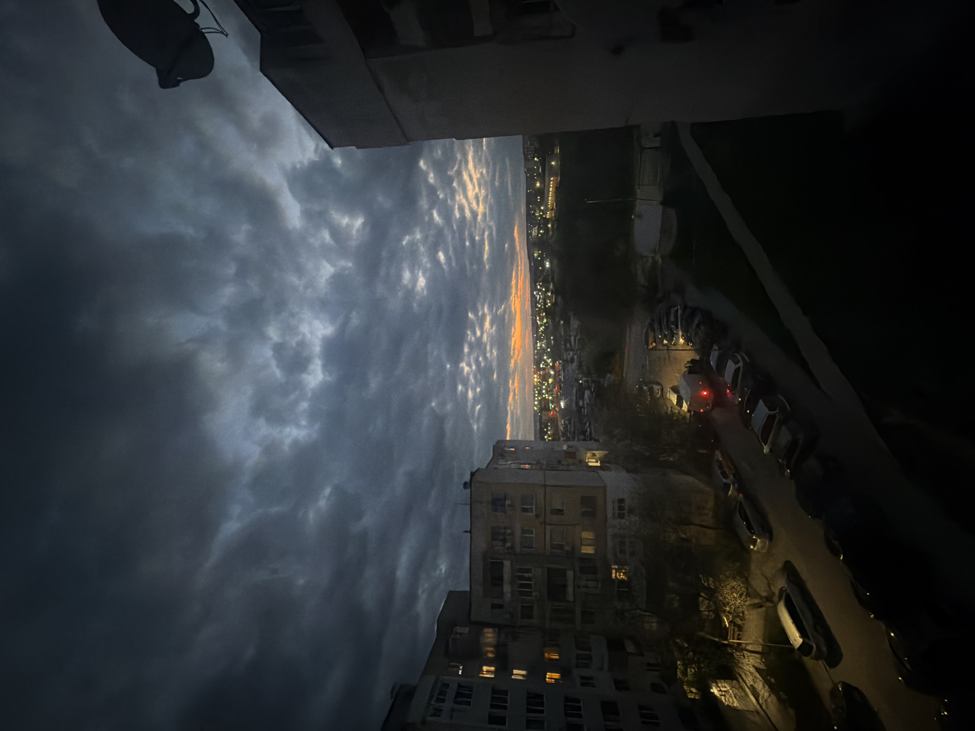 The image shows a nighttime view from an elevated position, likely a balcony or high-rise window, in Varna, Bulgaria. Dark, stormy clouds dominate the left side of the frame, contrasting with a thin strip of orange sky on the horizon, indicating either sunrise or sunset. In the distance, city lights are visible, suggesting an urban setting. Below, there are residential buildings, a street lined with parked cars, and a small patch of greenery. The lighting is dim, with artificial lights from buildings and streetlights illuminating the scene. The overall atmosphere is somber and urban. A satellite dish is visible on the top left corner.