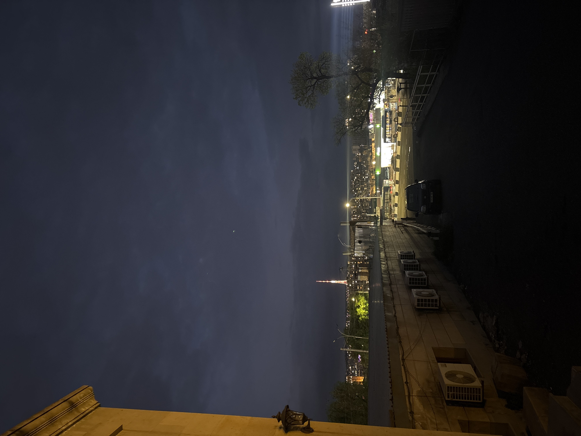 The image, correctly oriented, depicts an urban night scene in Yerevan, Armenia. The sky is a deep, dark blue-grey, suggesting twilight or early night, with faint clouds.
In the foreground, a warm, yellow-lit building facade or ledge occupies the bottom right, featuring a classic, ornate wall-mounted light fixture. On a lower roof or adjacent structure, a row of four white rectangular air conditioning units is visible, with another larger unit closer in the bottom right.
The midground reveals a city street illuminated by numerous streetlights, casting a cooler, white light. A dark-colored car, possibly a minivan or small SUV, is parked or driving on the road, with its front visible. Several trees with sparse foliage line the street, their forms distinct against the night sky. Buildings with numerous lit windows stretch into the distance, contributing to the urban glow.
In the background, against the dark horizon, a prominent and slender tower structure rises, illuminated with red lights. This is identifiable as the Yerevan TV Tower, serving as a key landmark in the cityscape. The overall scene is calm and quiet, characteristic of an urban environment after dark. No specific human activity is discernible, and no legible text is visible.