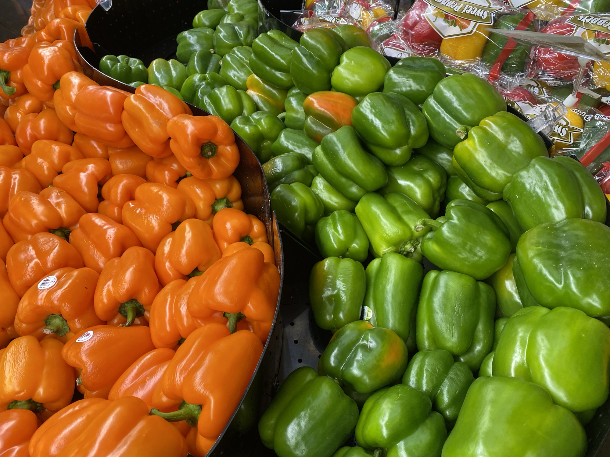 The image shows a display of orange and green bell peppers, likely in a grocery store or market setting. The peppers are densely packed, with the orange peppers on the left and the green peppers on the right. Some packaged bell peppers, including yellow and red varieties, are visible behind the green peppers. No people or identifiable landmarks indicating a specific location in San Bernardino Tlaxcalancingo, Mexico, are visible in the image.