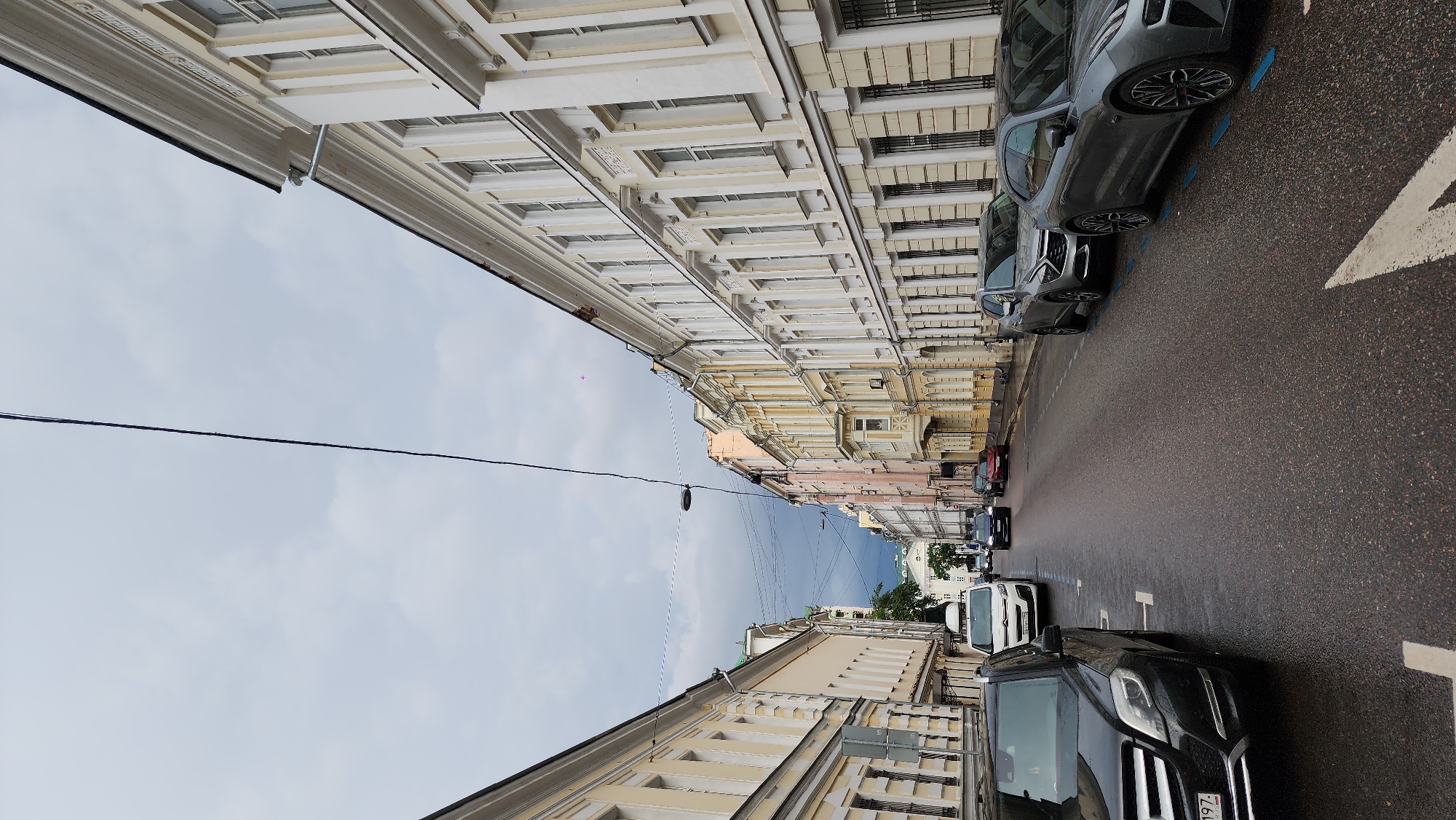 This is an eye-level, outdoor shot looking down a Moscow, Russia street. The street is narrow and lined with ornate buildings. The buildings are multi-storied and feature architectural details. Cars are parked along the right side of the street. A black car is parked on the right and a grey car is on the left, closer to the viewer. The street is wet. The sky is overcast. Powerlines run across the image.