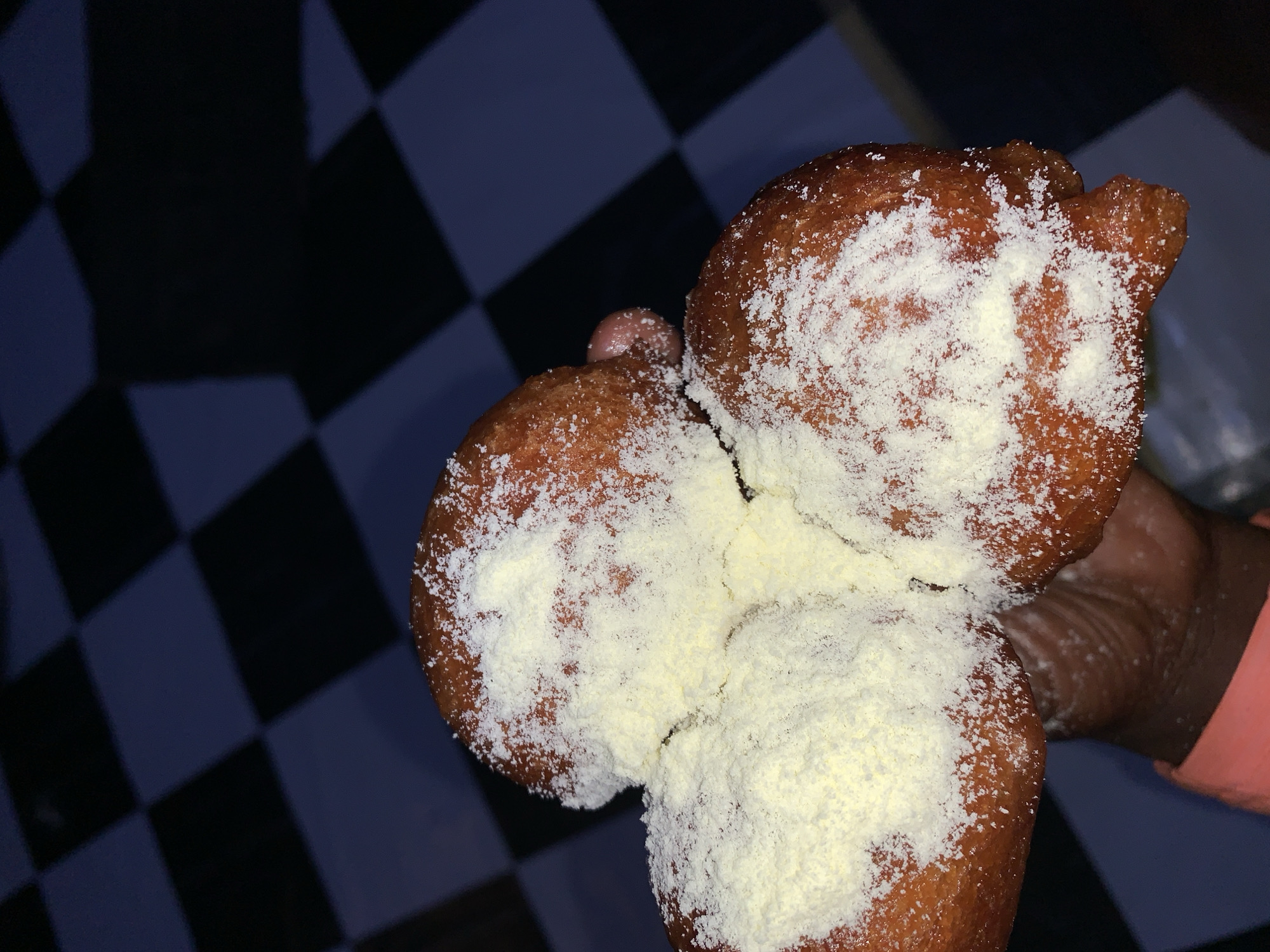A close-up shot shows three golden-brown fried dough balls lightly dusted with a white powder, possibly powdered sugar or cheese. The dough balls are clustered together in a person's hand, partially obscuring a pink sleeve. The background features a black and white checkered pattern, reminiscent of a floor or wall, with dark areas that could be shadows. The image appears to be taken in a casual indoor setting, possibly a home or a food stall, in Rafin Par, Nigeria.