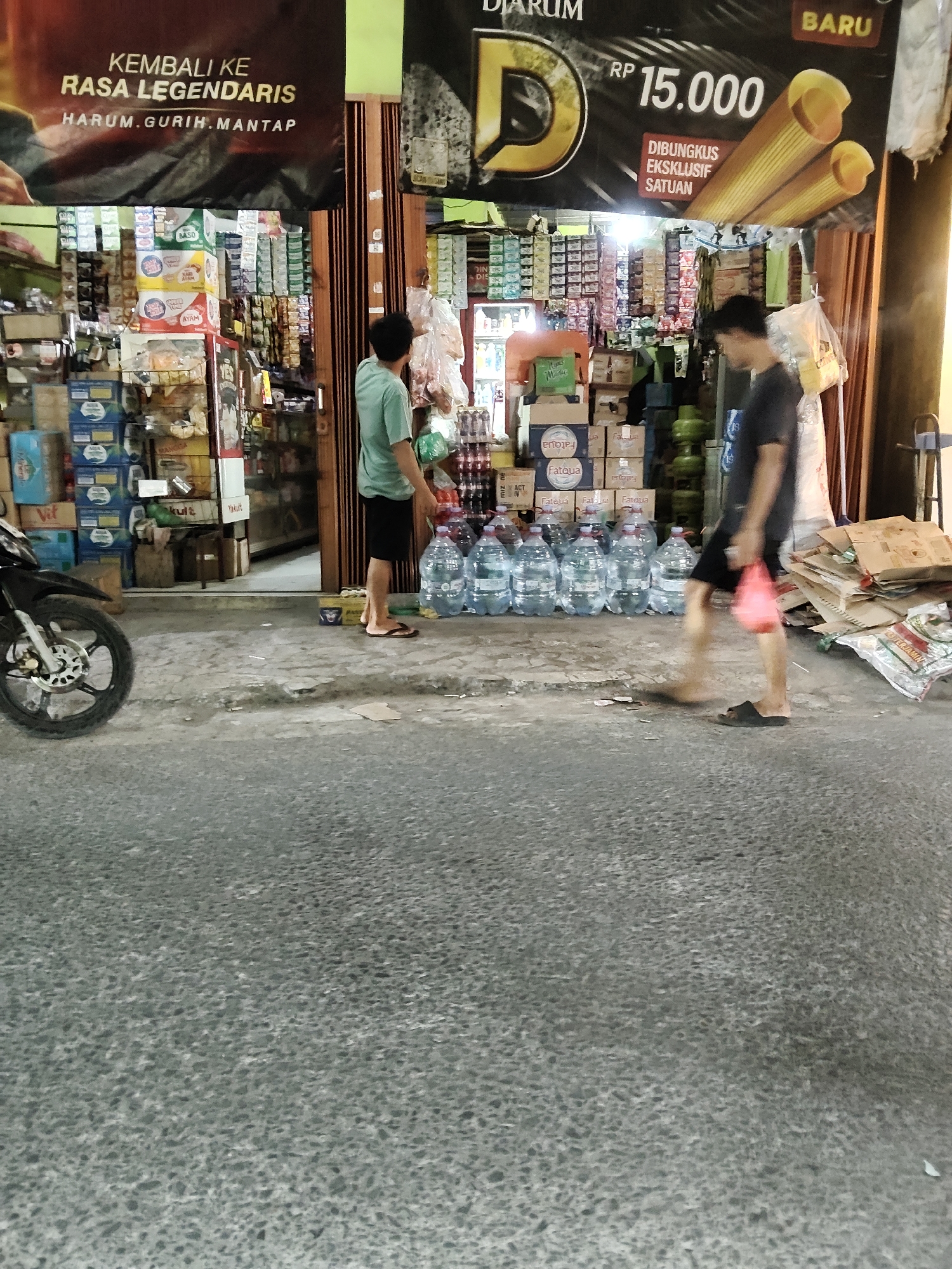 The image depicts a street scene in Mekarmukti, Indonesia, outside a small shop. Two men are present. One man, wearing a light green t-shirt and black shorts, stands facing away from the camera, near a display of bottled water. The second man, wearing a dark t-shirt and black shorts, walks away from the shop entrance, carrying a red plastic bag. In front of the shop, there are stacks of bottled water, various goods displayed inside, and a motorcycle parked on the left side of the frame. A large banner with text in Indonesian hangs above the shop. The ground is paved with asphalt and concrete. The environment appears to be an urban commercial area.