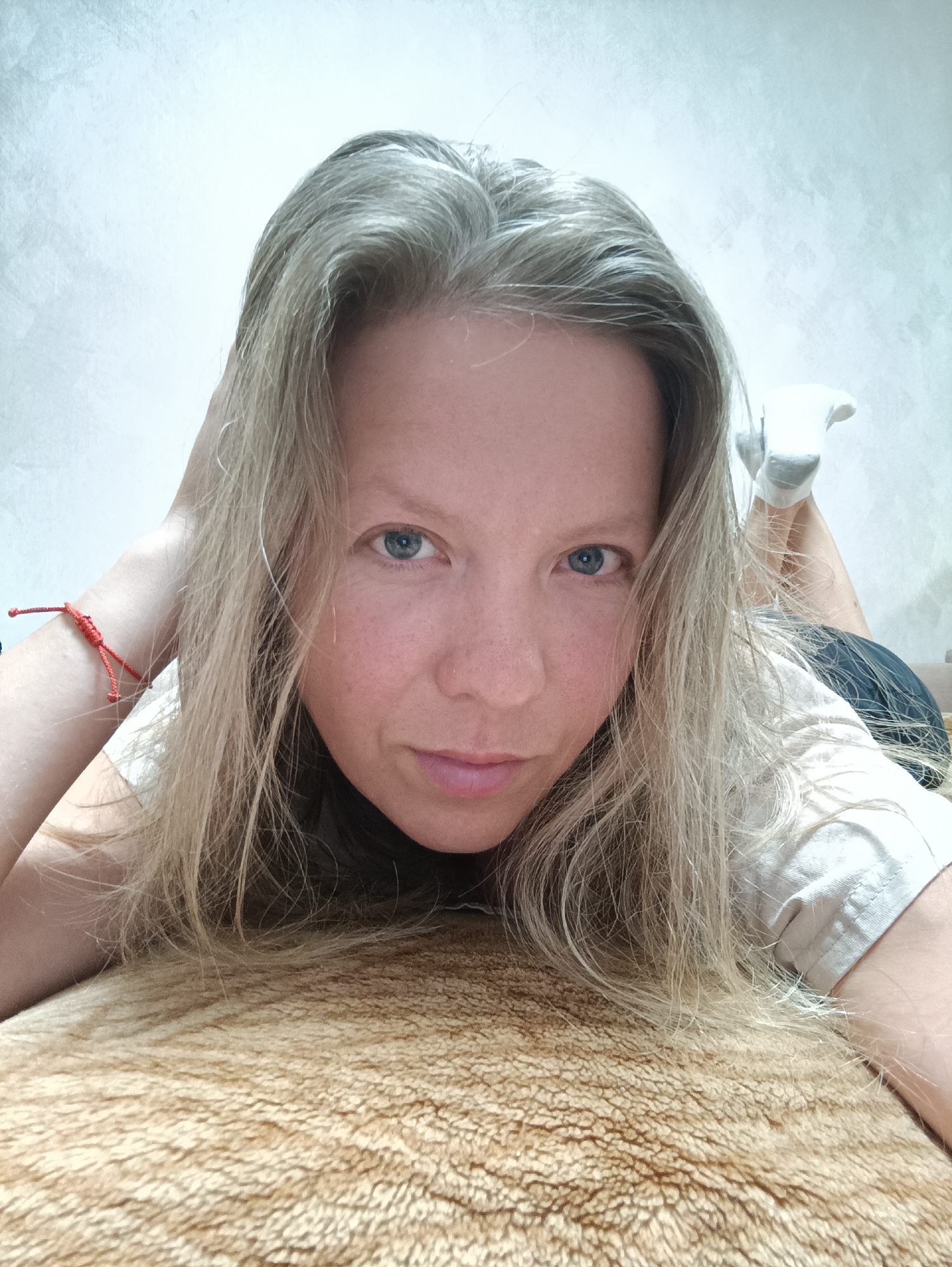 A woman with light blonde hair and blue eyes is lying on a tan-colored textured surface, possibly a bed or rug. She is wearing a light-colored t-shirt and a red bracelet on her left wrist. Her head is resting on the surface, and her right hand is holding her hair up. A pair of white socks is visible in the background. The background wall is plain and light-colored. The image appears to be a self-portrait. The city of Chaikovsky, Russia is not evident in the image.