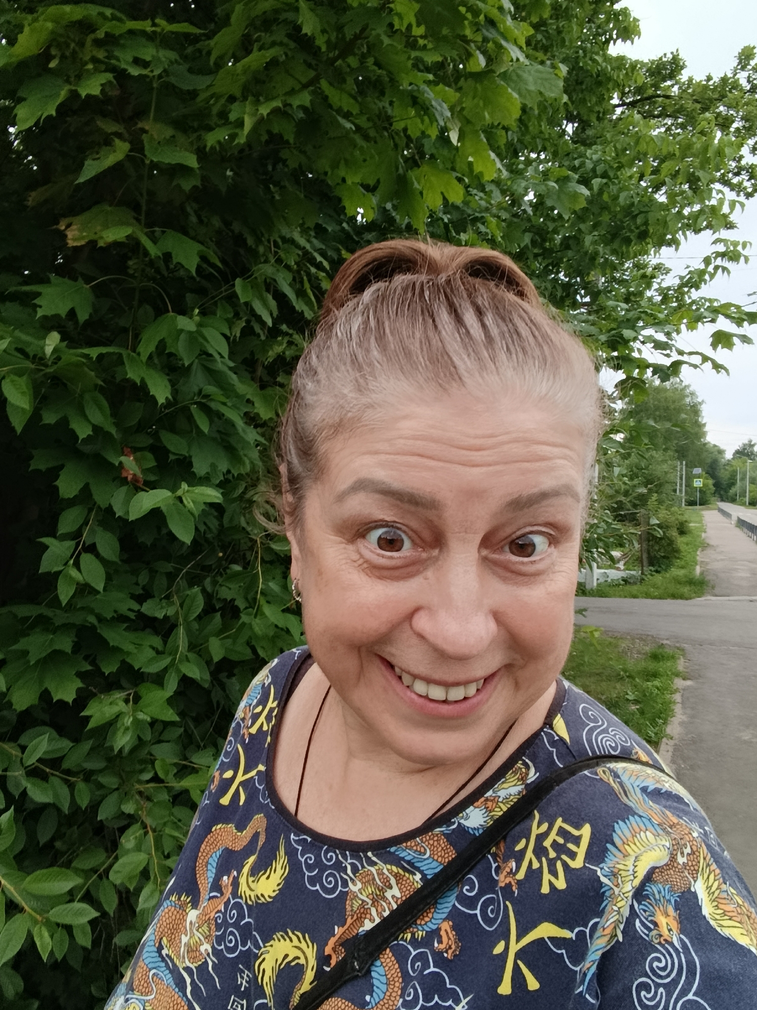 A woman with brown hair pulled into a bun and wide, excited eyes is taking a selfie outdoors. She is smiling broadly, showing her teeth. She is wearing a dark blue shirt with a repeating pattern of mythical creatures, possibly dragons and phoenixes, in yellow, orange, and blue. A thin black strap of a bag is visible across her shoulder.
The background consists of lush green foliage on the left, suggesting a wooded area or park. To the right, there is a paved path or road with a grassy verge. In the distance, a pedestrian crossing sign with an accompanying informational sign is visible. The overall impression is of a daytime scene, likely in a suburban or rural environment. The overcast sky indicates potentially cloudy weather. The woman's expression conveys amusement and a sense of spontaneity. There is some visible text on her shirt in yellow, appearing to be Asian characters, but it is not clearly legible. The location context provided is Верея, Russia.