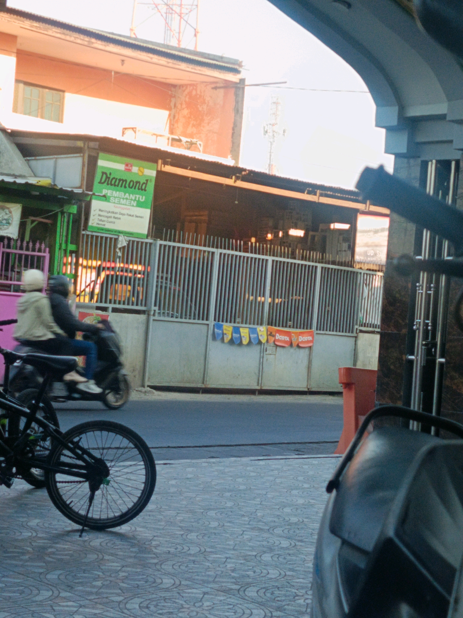 The image depicts a street scene in Kwangsan, Indonesia, bathed in the warm light of what appears to be either sunrise or sunset. In the foreground, a bicycle is parked on a patterned tiled sidewalk. Beyond the sidewalk, a street runs horizontally across the frame. On the opposite side of the street, a metal fence encloses a row of buildings. A person wearing a light-colored jacket and helmet is riding a motorcycle with a passenger behind them, moving away from the viewer. Above the buildings, a cell tower is visible against a bright sky. A green sign with "Diamond" prominently displayed hangs on one of the buildings. The overall impression is one of a typical Indonesian street scene during daylight hours, with elements of urban life and commerce.