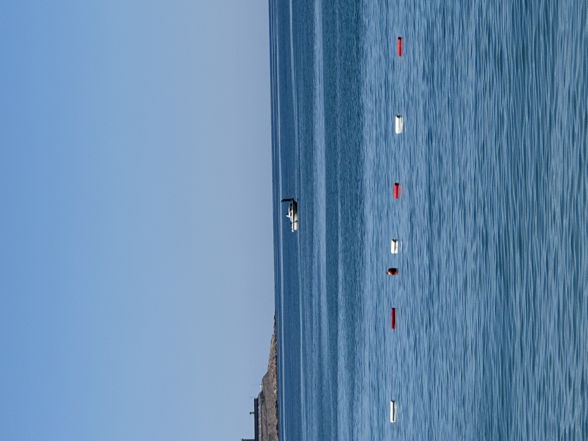 The image shows a body of water, possibly the Adriatic Sea, with a small boat and several red and white buoys floating on the surface. The sky is clear and blue. The location appears to be off the coast of Šušanj, Montenegro, based on the shoreline visible in the lower left of the image.