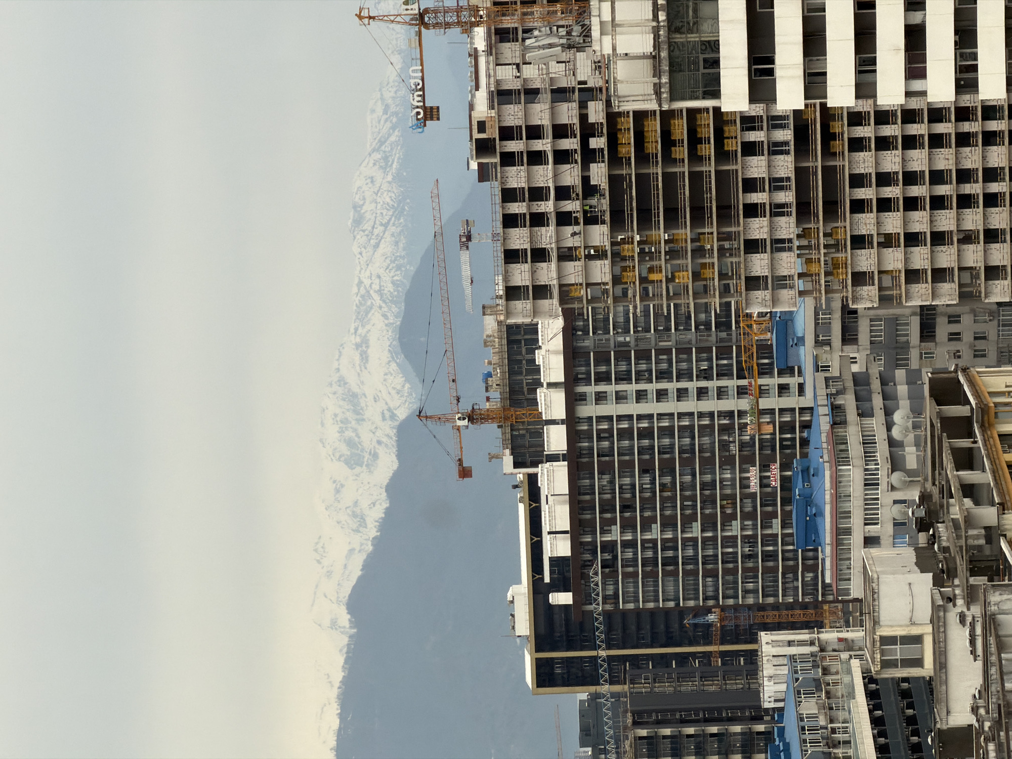 The image shows a construction site in Batumi, Georgia, with multiple high-rise buildings under construction and several tower cranes. The buildings are in various stages of completion, some with exposed frameworks. The environment includes mountains in the background, partially obscured by clouds.
