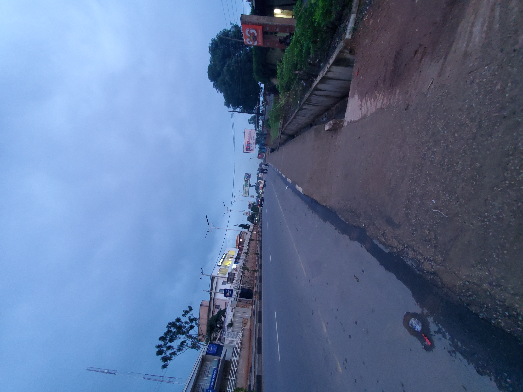 The image depicts a street scene in Jos, Nigeria, during twilight. A wide asphalt road dominates the foreground, with a distinct dark wet patch containing scattered debris including red and white plastic wrappers on the left side. A concrete sidewalk runs along the right side of the road, bordered by a concrete drainage channel. Beyond the sidewalk, lush green vegetation and a brick structure with an orange sign are visible.
On the left side of the road, buildings line the street. One building features a white facade with blue signage that reads "Colour Centre." Another building has a multi-story structure with balconies and white railings. Several streetlights are visible along the road.
Further down the road, the sky is a pale blue with scattered clouds. In the distance, several vehicles, including cars and what appear to be vans, are driving on the road. Billboards are also present along the roadside. The overall impression is a moderately busy urban street at the end of the day.