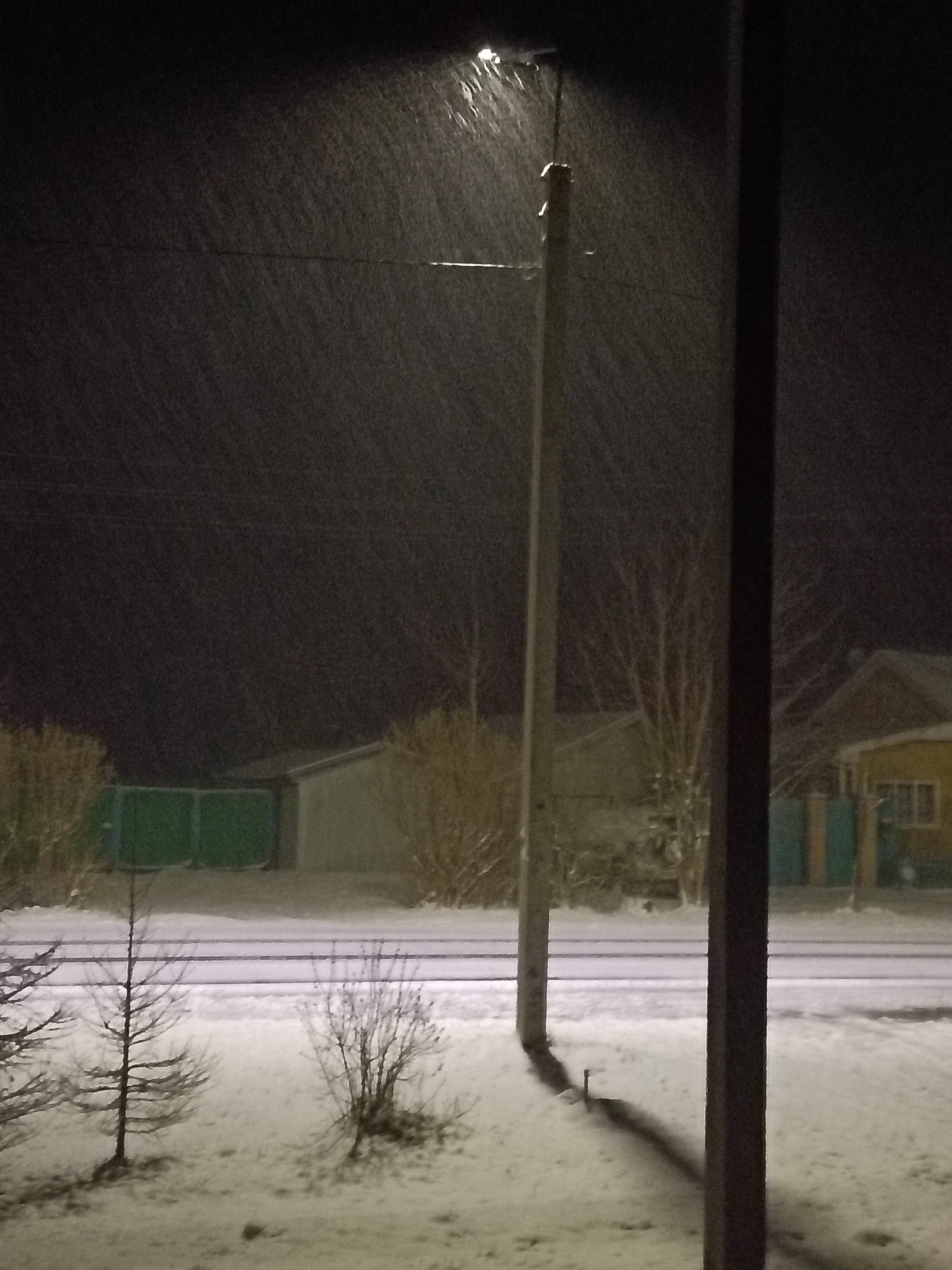 The image shows a snowy night scene in what appears to be a residential area of Красный Маяк, Russia. A street lamp illuminates the falling snow. In the background are several buildings, including a shed with a green gate and a yellow house. A power line is visible connecting to the street lamp. In the foreground, snow covers the ground, and several bare trees and shrubs are present. Two vertical posts are visible in the foreground. The scene is quiet and dark.