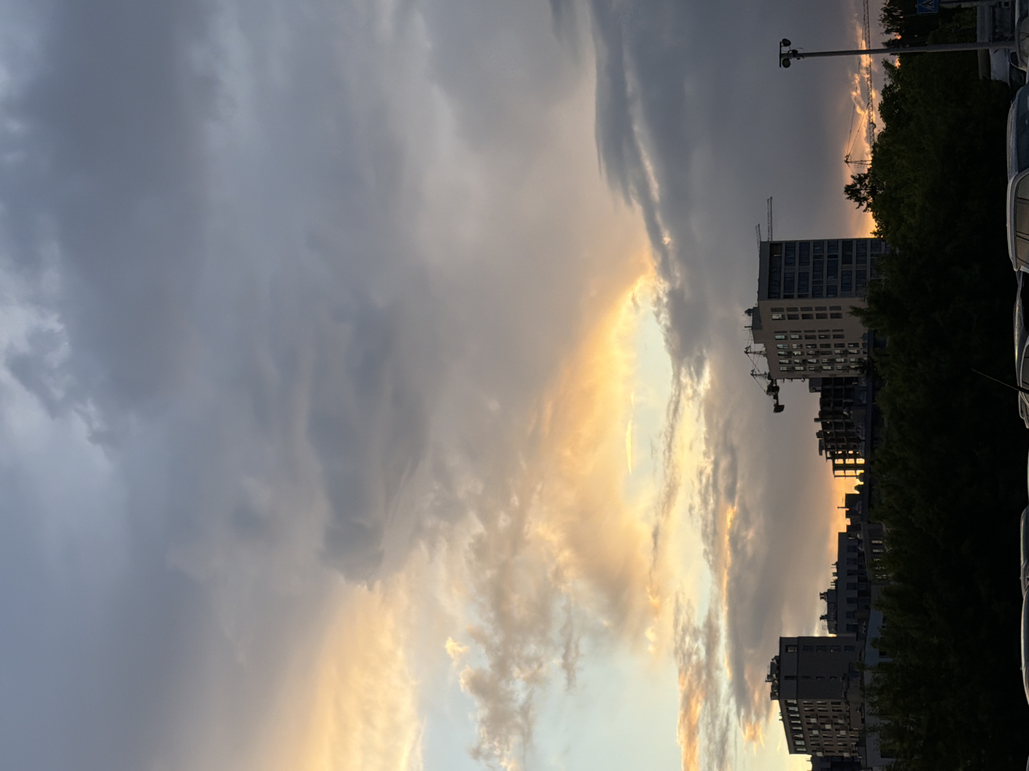 The image depicts a cityscape at dusk with dramatic cloud formations. In the foreground, heavy, dark clouds dominate the left side of the frame, contrasting with the lighter, golden-hued clouds toward the center. Buildings are silhouetted against the bright sky. A crane and a few buildings under construction are visible. There are some trees visible between the buildings. Cars are parked on the right. The city of Izhevsk, Russia, is not discernible from the image.