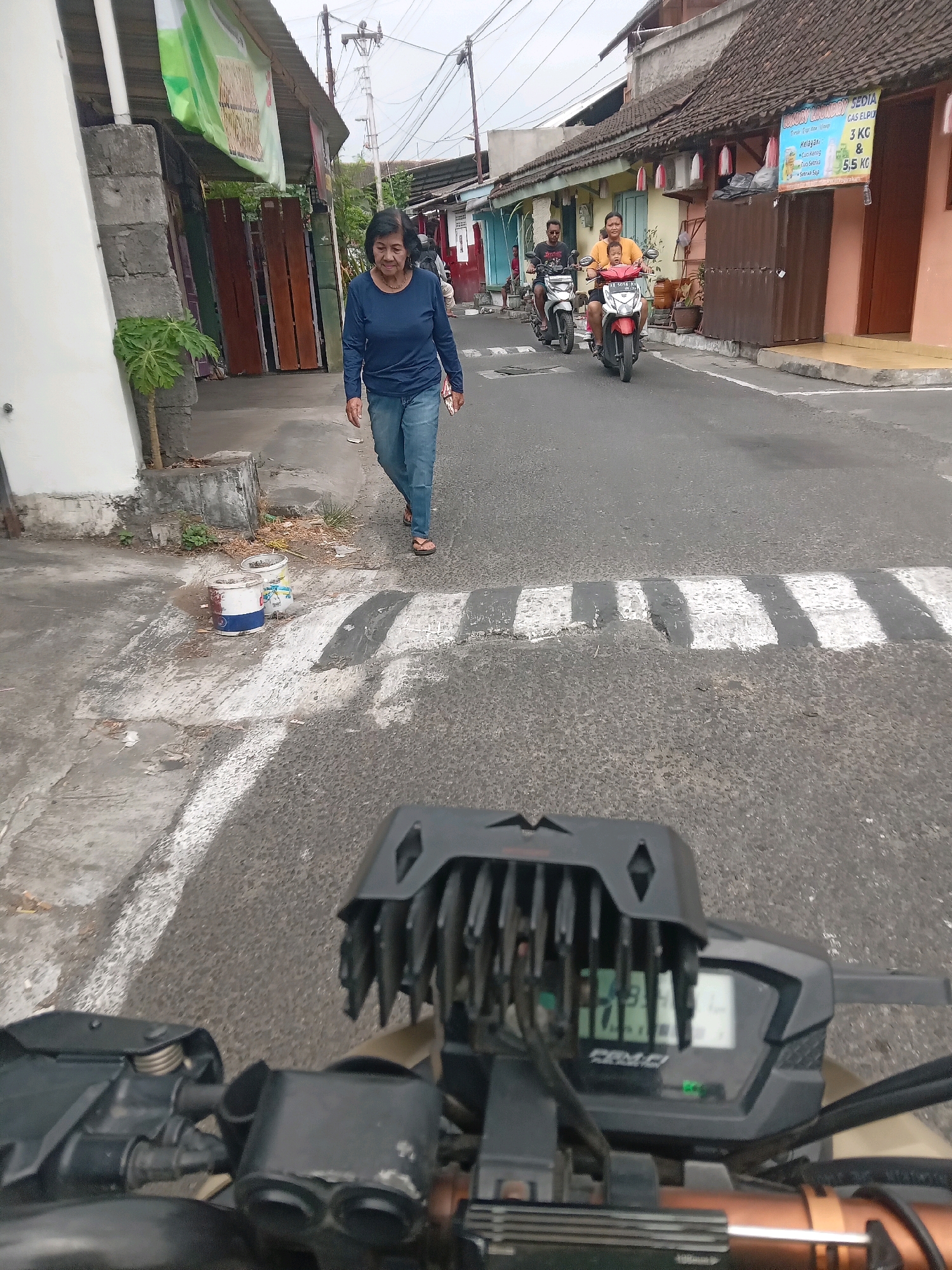 The image depicts a street scene in Yogyakarta, Indonesia, likely in a residential or commercial area. In the foreground, a motorcycle's handlebars and dashboard are visible, indicating the photo was taken from the rider's perspective. The dashboard shows a digital display with various indicators.
Further down the street, an elderly woman wearing a blue long-sleeved shirt and jeans walks on the sidewalk. She is looking to her right. Two people on motorcycles are riding in the street. The first motorcycle, a white scooter, has a man riding it, and the second, a red scooter, has a person in an orange shirt riding it, with another person possibly behind them or obscured.
The street itself is paved and has white painted markings, possibly indicating a pedestrian crossing or speed bump. Several buildings line the street, some with tiled roofs and painted facades. One building on the right has a sign with text that appears to be in Indonesian, advertising laundry services.
On the sidewalk near the woman, there are two buckets, one white with red and blue markings, and another white bucket. Small plants are visible in pots or planters along the sidewalk. Electrical poles and wires are present in the background, typical of an urban environment. The overall lighting suggests it is daytime, possibly overcast.