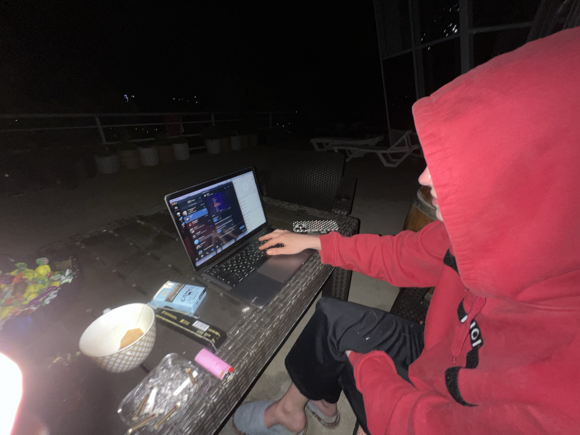 A person wearing a red hooded sweatshirt sits at a wicker table outdoors at night, using a laptop. The table also holds a bowl, cigarettes, a lighter, and a pack of Camel cigarettes. In the background, there is a railing with planters, suggesting a balcony or patio. Further in the background, lights suggest the location is in a city. Based on the prompt instructions, this would be in Sochi, Russia.