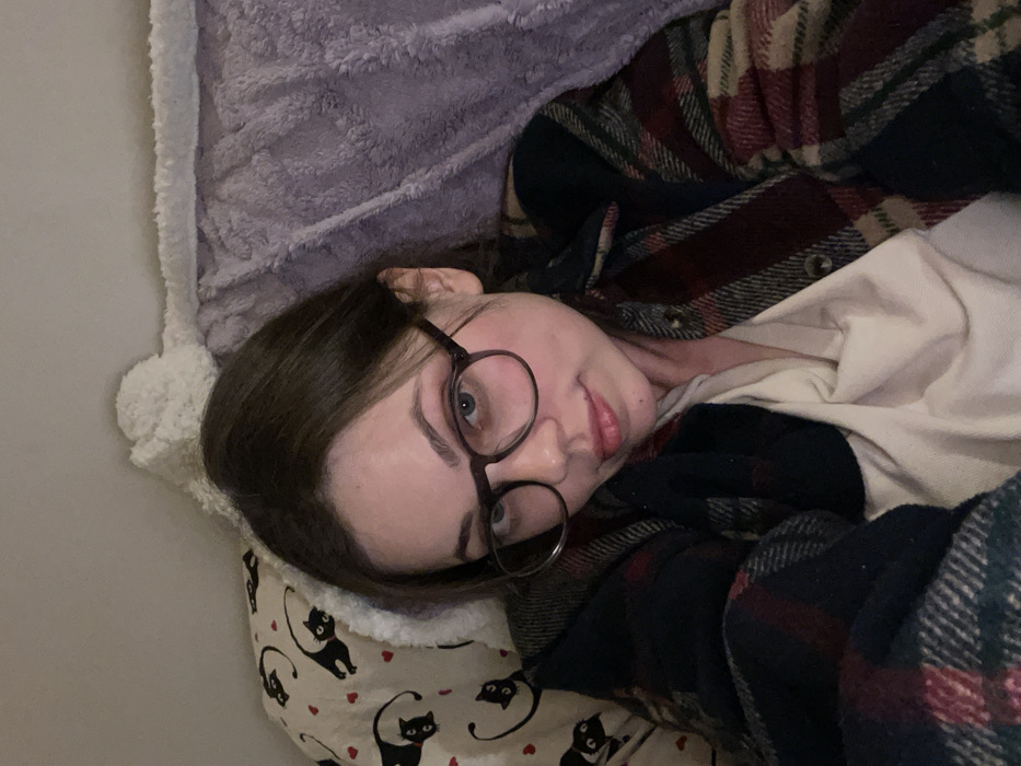 A female individual is depicted in a close-up, indoor setting within Monsummano Terme, Italy. She possesses dark brown hair and wears dark-rimmed circular eyeglasses, looking directly into the camera. Her expression is neutral to slightly smiling, with lips slightly parted. She is dressed in a plaid flannel shirt, displaying colors including dark blue/green, maroon, and beige, worn over an off-white undergarment. The subject appears to be in a reclining or lying posture. To the left side of the frame (viewer's right), a textile object, possibly a pillow or blanket, features a pattern of black cats and small red hearts on a light background. To the right side of the frame (viewer's left), a plush, light purple blanket is visible. A white, textured, fluffy object is positioned behind the subject's head. The background consists of a plain, light-colored wall. The scene is illuminated by soft, ambient indoor lighting.