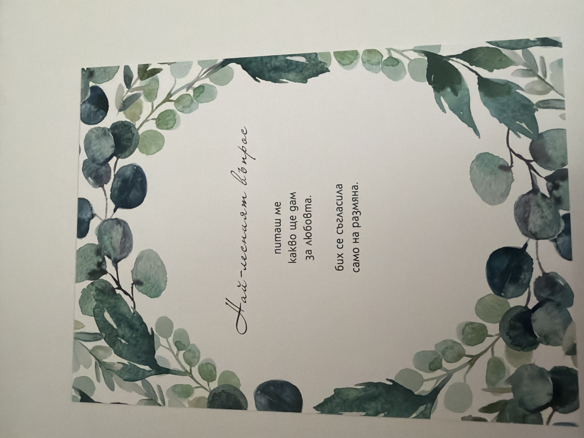 The image is a close-up shot of a card or invitation with a watercolor floral border, featuring eucalyptus leaves and branches in various shades of green and blue. The text is written in Bulgarian and is positioned centrally within the floral design. The overall aesthetic is delicate and natural. There is no information in the image to suggest a location in Pazardzhik, Bulgaria, or any other specific city. The image depicts a stationary item and contains no people, actions, or interactions.
