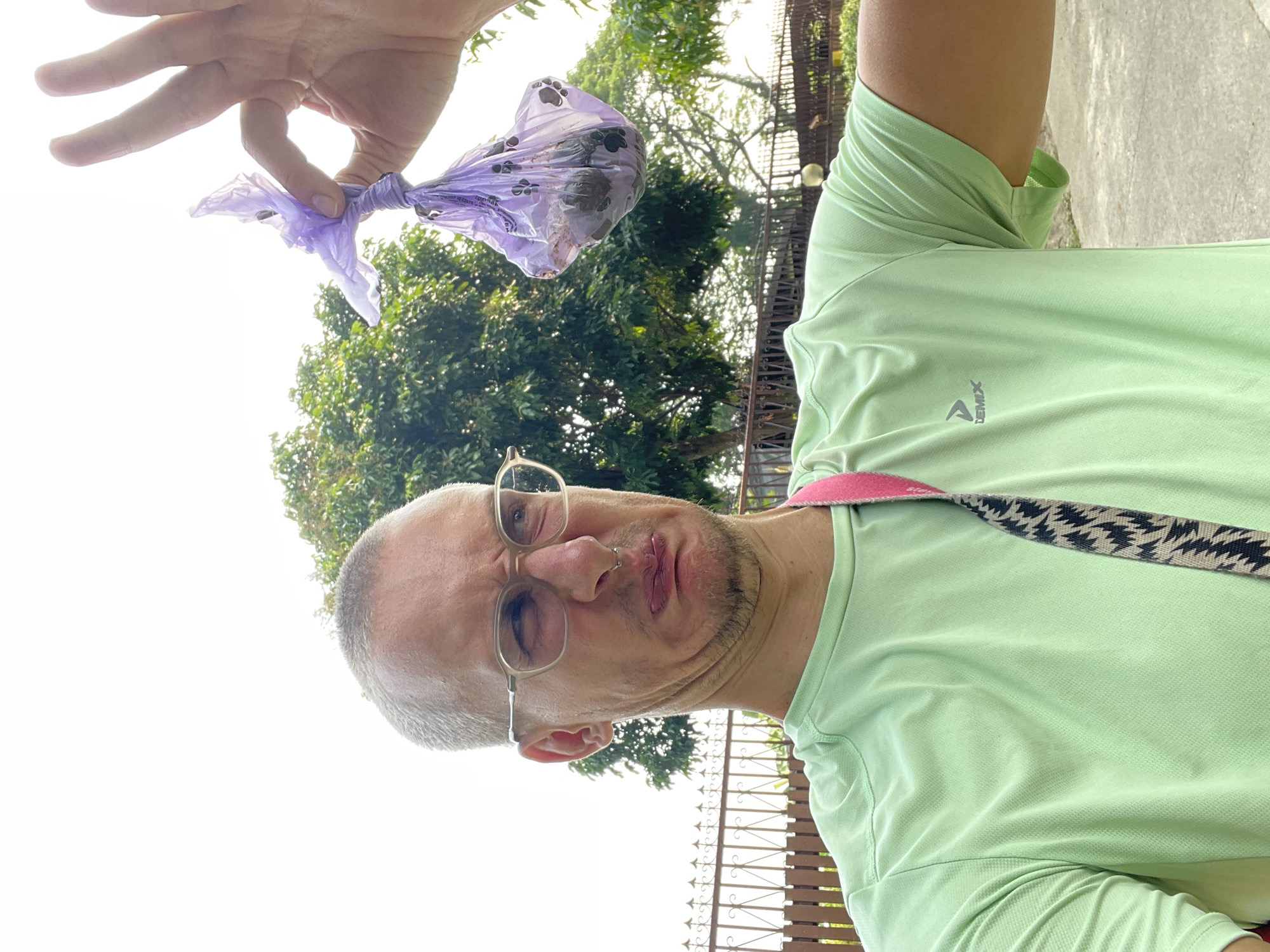 A light-skinned man with short, gray hair and glasses holds up a purple plastic bag containing dog feces. The man wears a light green athletic shirt and a patterned strap across his chest. He makes a disgusted facial expression. Behind the man is green foliage, a fence, and the sky. The scene appears to be outdoors, and the context suggests the location is Fa Ham, Thailand.