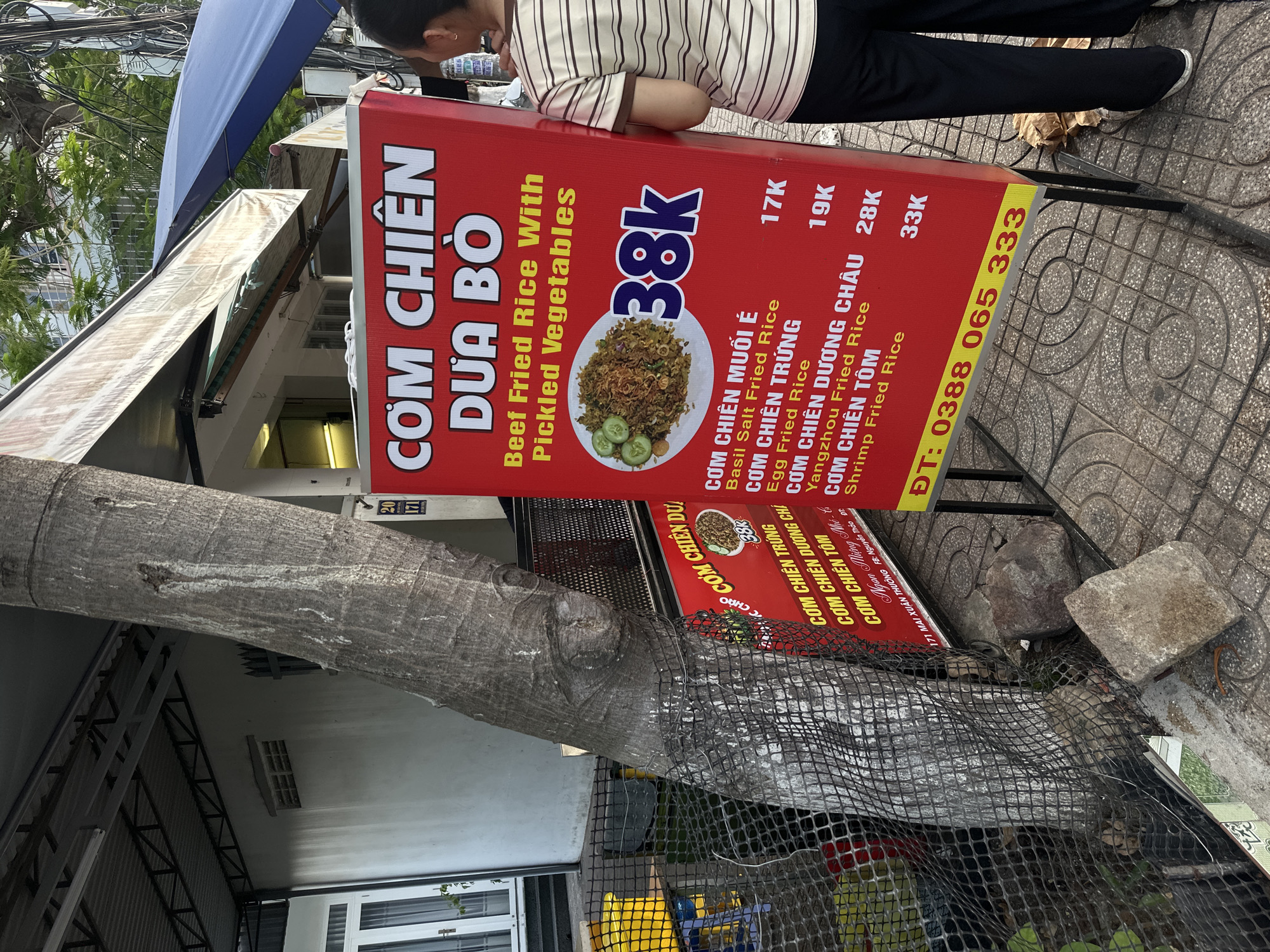 A red sign with Vietnamese text and English translations advertises fried rice dishes. The sign is placed on a paved sidewalk next to a tree and a black mesh fence. A person wearing a striped shirt and dark pants stands to the right of the sign, facing away from the camera. In the background, a building with a corrugated metal roof and white walls is visible. The scene appears to be a street food stall in Bac Nha Trang Ward, Vietnam.