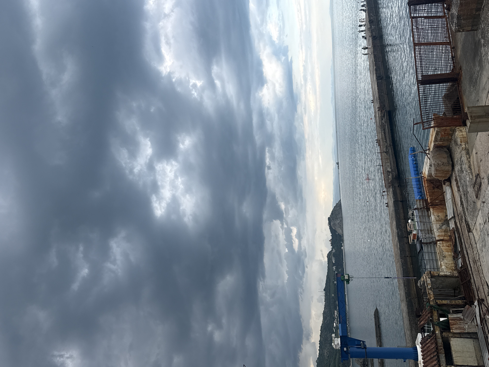 The image captures a view of a harbor with a cloudy sky. The perspective is from a slightly elevated position, looking out towards the water.
Dominating the left side of the frame are dark, heavy clouds that suggest an overcast or potentially stormy day. These clouds create a dramatic backdrop. On the right, the sky transitions to brighter, lighter tones, with glimpses of sunlight breaking through. The sea is visible, appearing calm with gentle ripples.
In the foreground on the right, there is a weathered concrete structure with visible signs of disrepair, including rusted metal elements and deteriorating walls. Adjacent to this structure, a blue crane with a long arm extends out over the water. Along the edge of the harbor, a concrete pier runs parallel to the water, with several moored boats or structures visible in the distance. Further out, a hilly coastline with some buildings is discernible against the lighter sky.
The overall mood is somewhat somber due to the heavy clouds, but the presence of the sea and the hint of brighter sky on the horizon offer a sense of expanse. The time of day appears to be daytime, possibly late afternoon or early evening, given the quality of the light and the cloud cover. There is no visible text in the image.