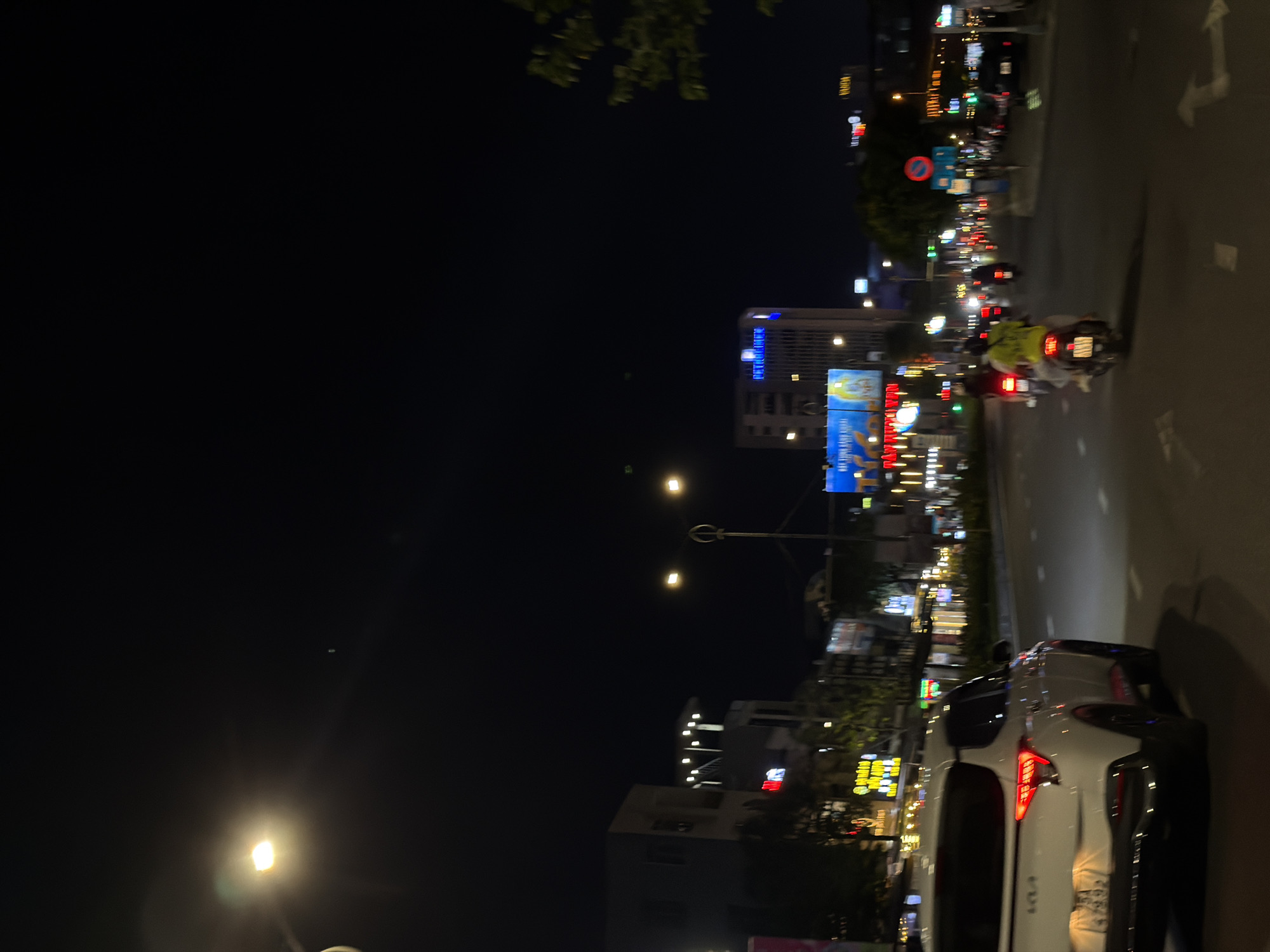 This media file depicts a nighttime scene on a street in Đà Nẵng, Vietnam. The perspective is from the side of a white car, looking towards the street and surrounding buildings. The image is oriented vertically, with the street running diagonally from the bottom right to the top left.
The primary subjects are a white car in the foreground, a scooter with two people riding it in the middle ground, and various buildings with illuminated signs and lights in the background. The scooter rider in front is wearing a light-colored jacket, and the passenger behind is wearing a bright green jacket. The car's taillights are illuminated, suggesting it is either stopped or moving slowly.
The scene is set at night, with the dark sky contrasting with the vibrant lights from businesses, streetlights, and vehicle lights. The weather appears to be clear, as there are no indications of rain or fog. The presence of multiple scooters and the style of the buildings and signage suggest an urban environment in Southeast Asia, specifically Đà Nẵng as indicated by the location context.
Notable details include the bright, colorful neon signs on the buildings, which are characteristic of many Asian cities. There is also a traffic sign with a red circle and a diagonal line, indicating a prohibition. The street is paved and appears to be well-lit. The overall mood is one of a bustling city at night, with a sense of movement and activity.
Visible text includes various signs on the buildings, some of which are in Vietnamese. For example, on a large billboard in the mid-ground, there appears to be text in Vietnamese, likely advertising a product or service. The car in the foreground also has some visible text on its rear, though it is partially obscured.