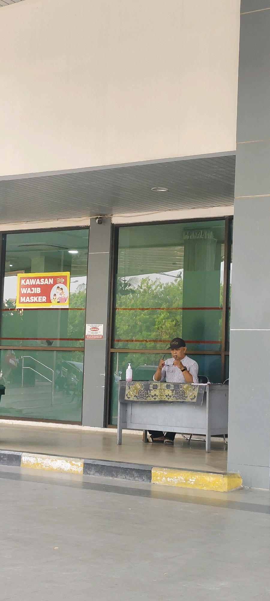 The image shows a man sitting at a desk outside a building. He is wearing a striped shirt, dark pants, and a cap. He is holding a pair of glasses to his face. On the desk, there is a bottle of hand sanitizer and a decorative cloth. Behind him, there are large glass windows that reflect the trees and sky outside. To the left of the man, there is a sign that says "KAWASAN WAJIB MASKER," which translates to "Mandatory Mask Area." The image appears to have been taken in a public area, possibly an entrance to a building, in Banjarmasin, Indonesia.