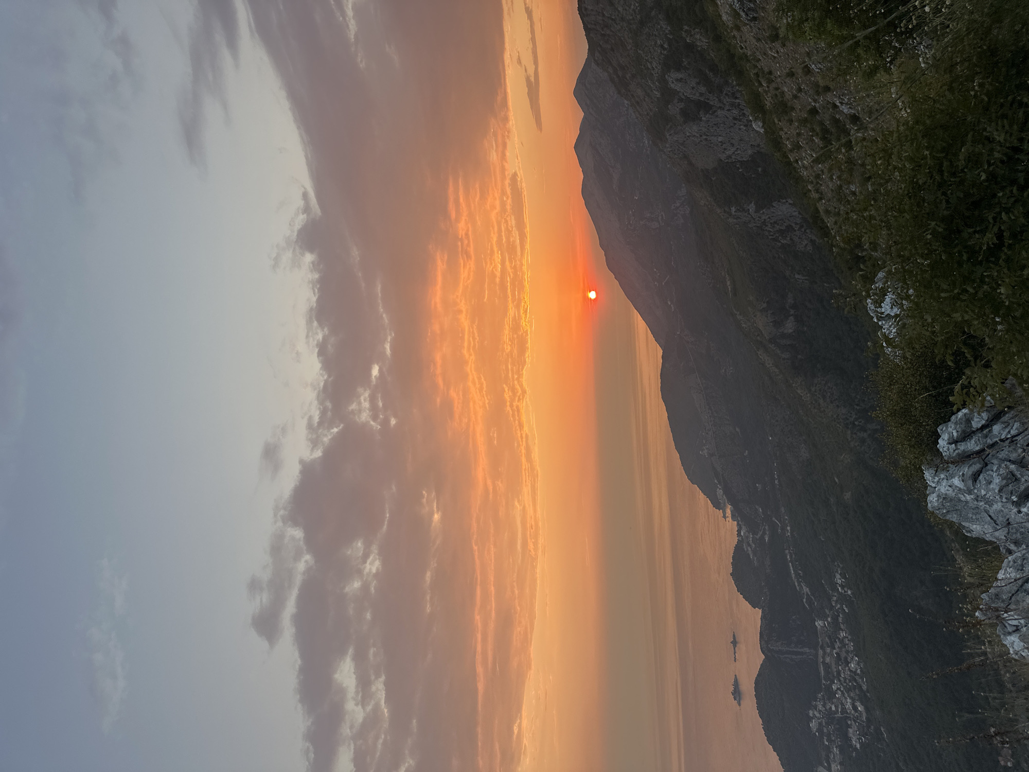 The image depicts a serene sunset over the Adriatic Sea, viewed from the mountainous terrain near Gluhi Do, Montenegro. The sky is a gradient of soft blues and oranges, with scattered clouds catching the last rays of the sun. The sun itself, a vibrant orb, is partially obscured by a mountain range in the mid-ground.
The landscape features rolling hills covered in dense green vegetation, interspersed with rocky outcrops. In the distance, the coastline is visible, with a small settlement nestled along the hillsides. The calm sea reflects the warm hues of the sky. The foreground is dominated by a rough, grey rock formation and some dry grasses. The overall atmosphere is tranquil and picturesque.