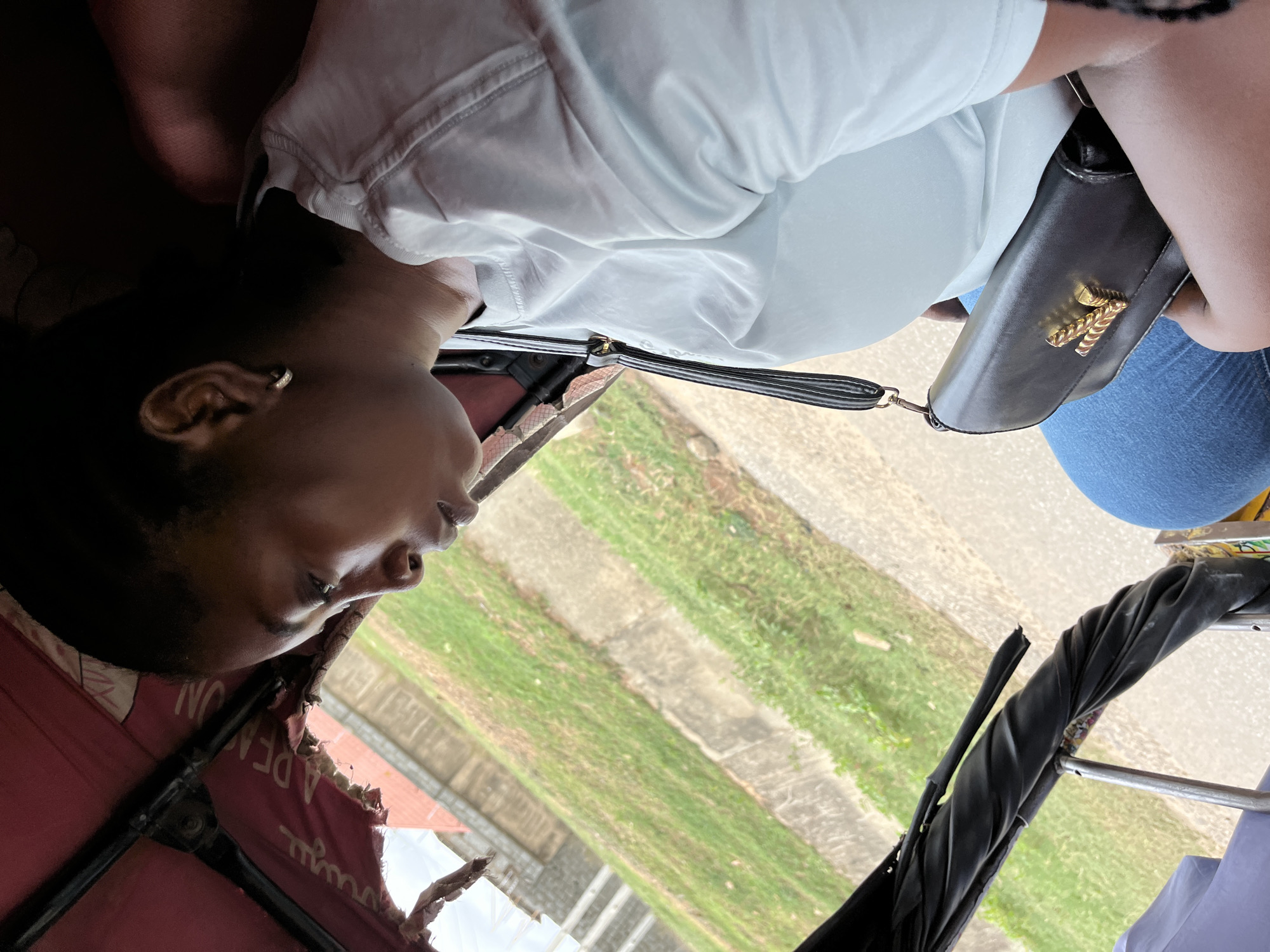 A photograph taken in what appears to be a motorized tricycle, commonly known as a "Keke Napep" in Nigeria. An individual is visible, likely a passenger, wearing a light-colored top. They are seated in the vehicle, and their facial features are partially visible. A black purse with a gold emblem is positioned near the passenger. The background features typical roadside scenery, with glimpses of greenery and a concrete sidewalk. Based on the presence of Keke Napeps and general appearance, the location is inferred to be in Abuja, Nigeria. The scene seems to be a normal snapshot of everyday life in the city.