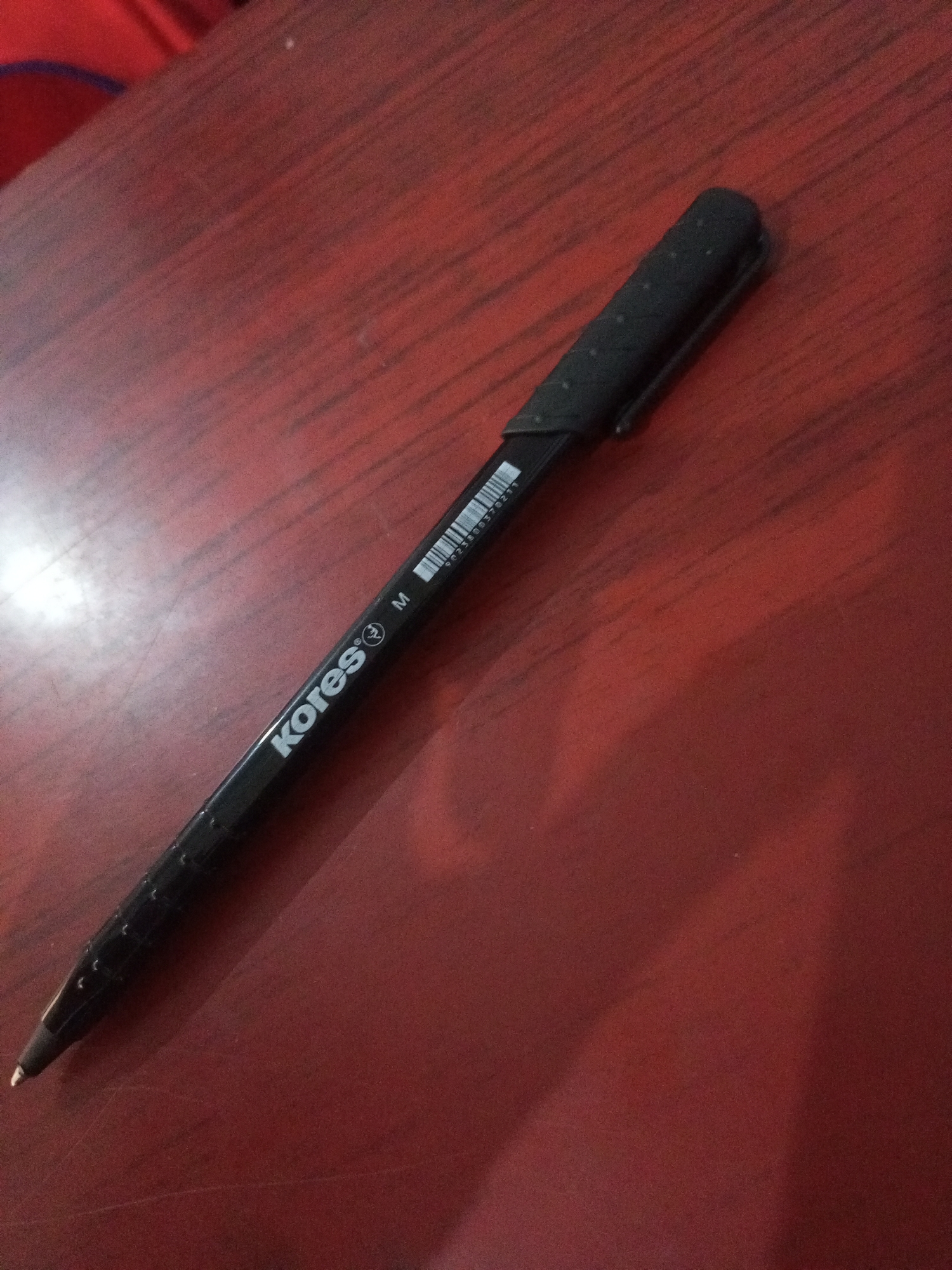 The image shows a black pen lying on a red, wooden surface. The pen has white lettering that says "kores" and the letter "M". There is also a barcode on the pen. The surface appears to be a table or desk. The lighting suggests an indoor setting, possibly a home or office. There are no people or other objects visible in the immediate vicinity of the pen. The image does not contain any information about the location in Santa Ana, Venezuela.