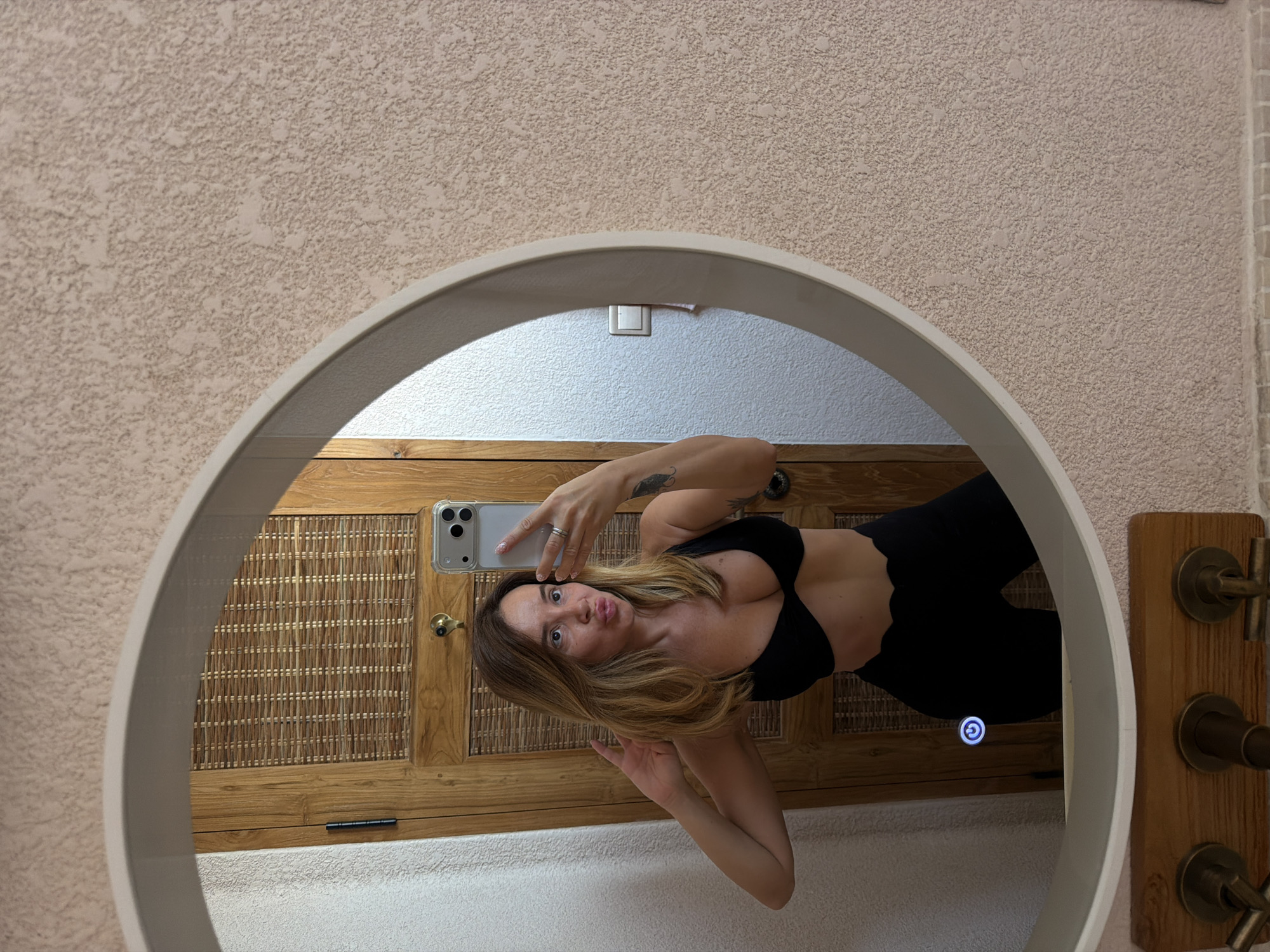 A woman is taking a selfie in a mirror. She is wearing a black bikini top and black leggings. The mirror is oval and has a lighted border. Behind her, a wooden door with woven panels can be seen. The walls are a textured beige color. The scene is likely a bathroom or dressing area. The provided crops hint at details of the woman's appearance, the phone she is holding, and the textures of the environment. Specific location in Mengwi, Indonesia is not determinable from the image alone.