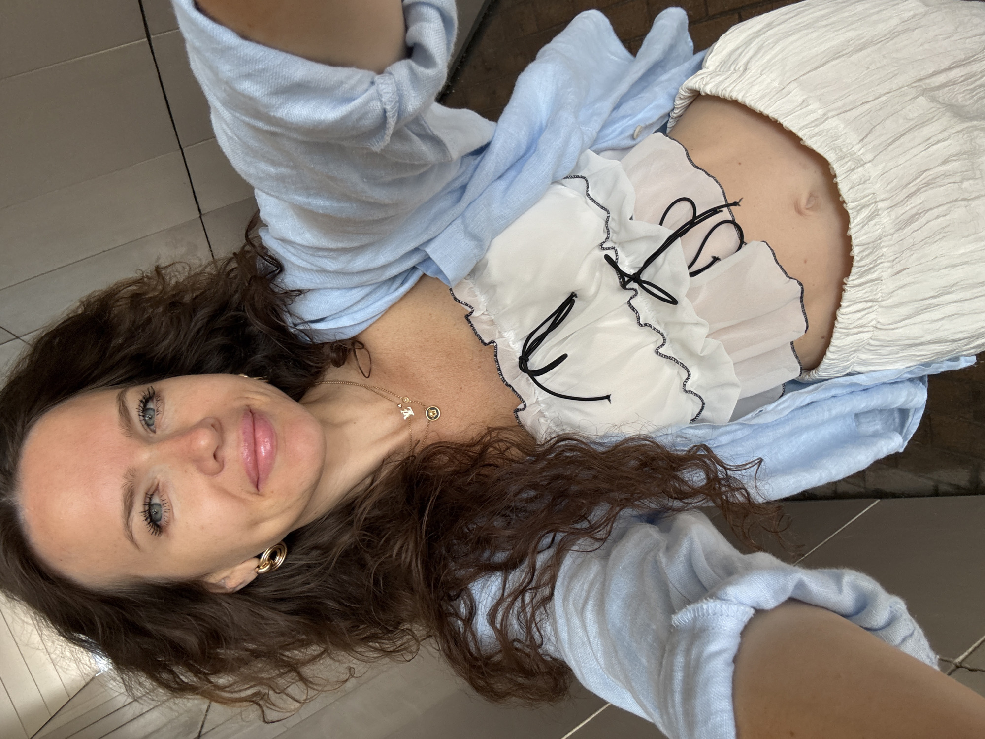 A woman is taking a selfie in a city environment. She is standing and her torso and head are visible in the frame. She has long, dark, curly hair and is wearing a light blue shirt, a white top with black trim, and white pants. She is also wearing gold earrings and a necklace. The background is blurred and appears to be an indoor setting with tiled walls. The location is identified as Khimki, Russia.