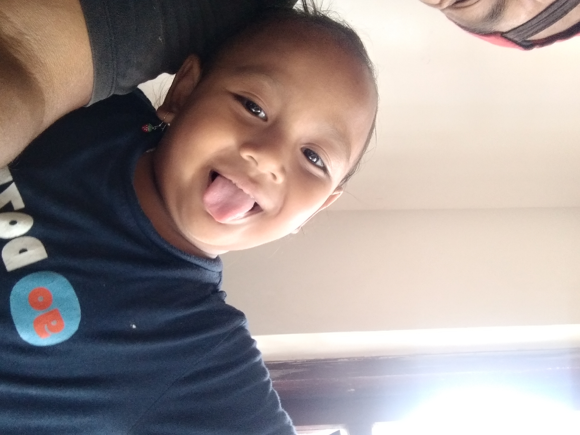 In a close-up, indoor selfie, a young child, likely of Southeast Asian descent, sticks their tongue out playfully. The child's head is tilted to the left, with dark hair visible. They are wearing a dark blue t-shirt with a graphic logo featuring an orange oval with red lettering. A small strawberry-shaped earring dangles from their left ear. To the left of the child, an adult's head and shoulder are partially visible, wearing a dark t-shirt and a red and black headband. The background consists of a plain, light-colored wall and a bright, sunlit window. The image appears to have been taken in the city of Pandak, Indonesia.