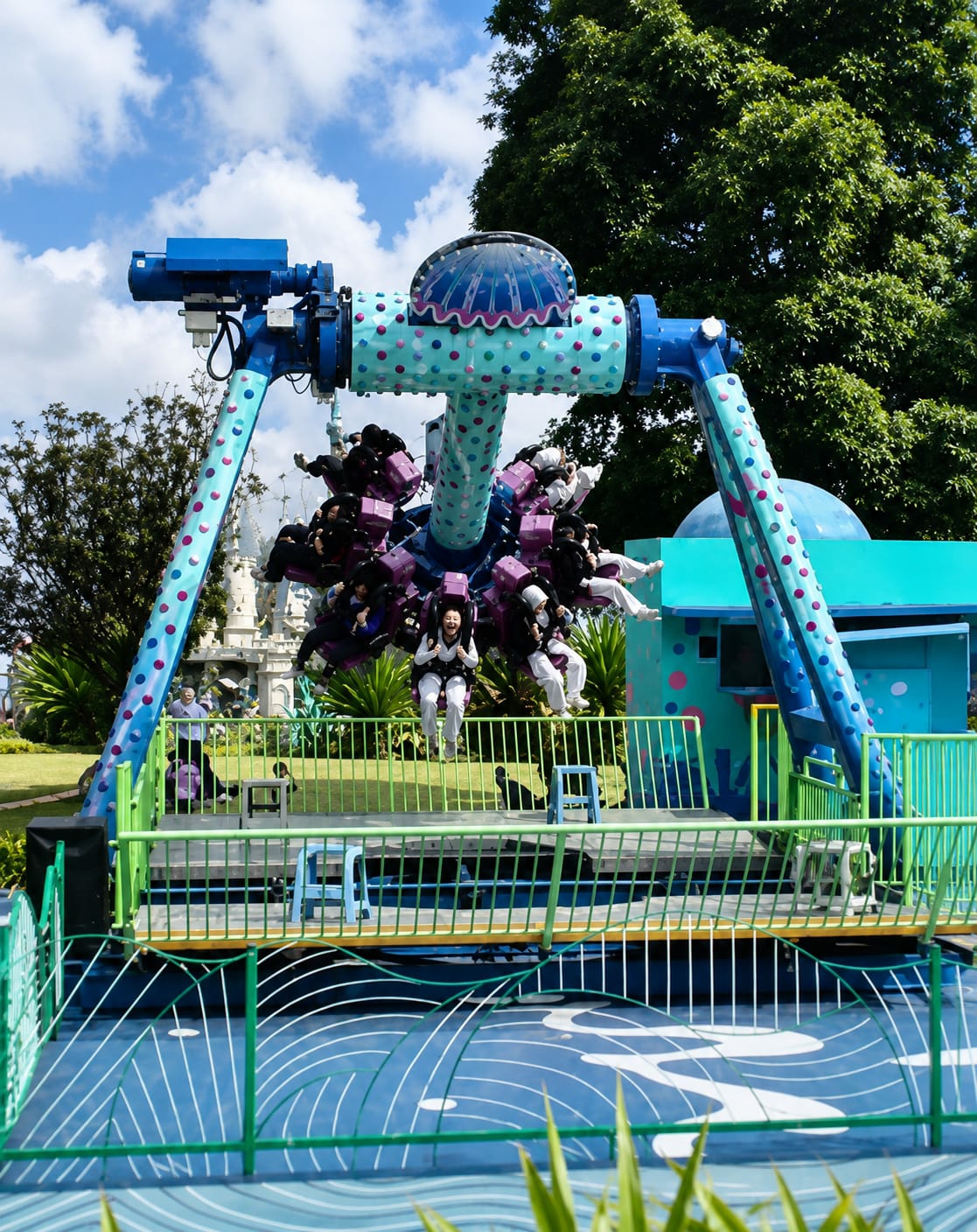 This image captures an amusement park ride in Berastagi, Indonesia, on a sunny day with a partly cloudy sky. The ride, a pendulum-style swing, is in motion, with several passengers seated in suspended carts. The ride's structure is primarily blue and teal, decorated with pink and purple polka dots. The passengers, dressed in casual attire and some in white uniforms, appear to be experiencing excitement, with one individual in the foreground smiling broadly. The ride is set against a backdrop of lush green trees and a light blue building with playful polka dot decorations. In the foreground, a blue and green safety barrier encloses a blue patterned surface that resembles water.