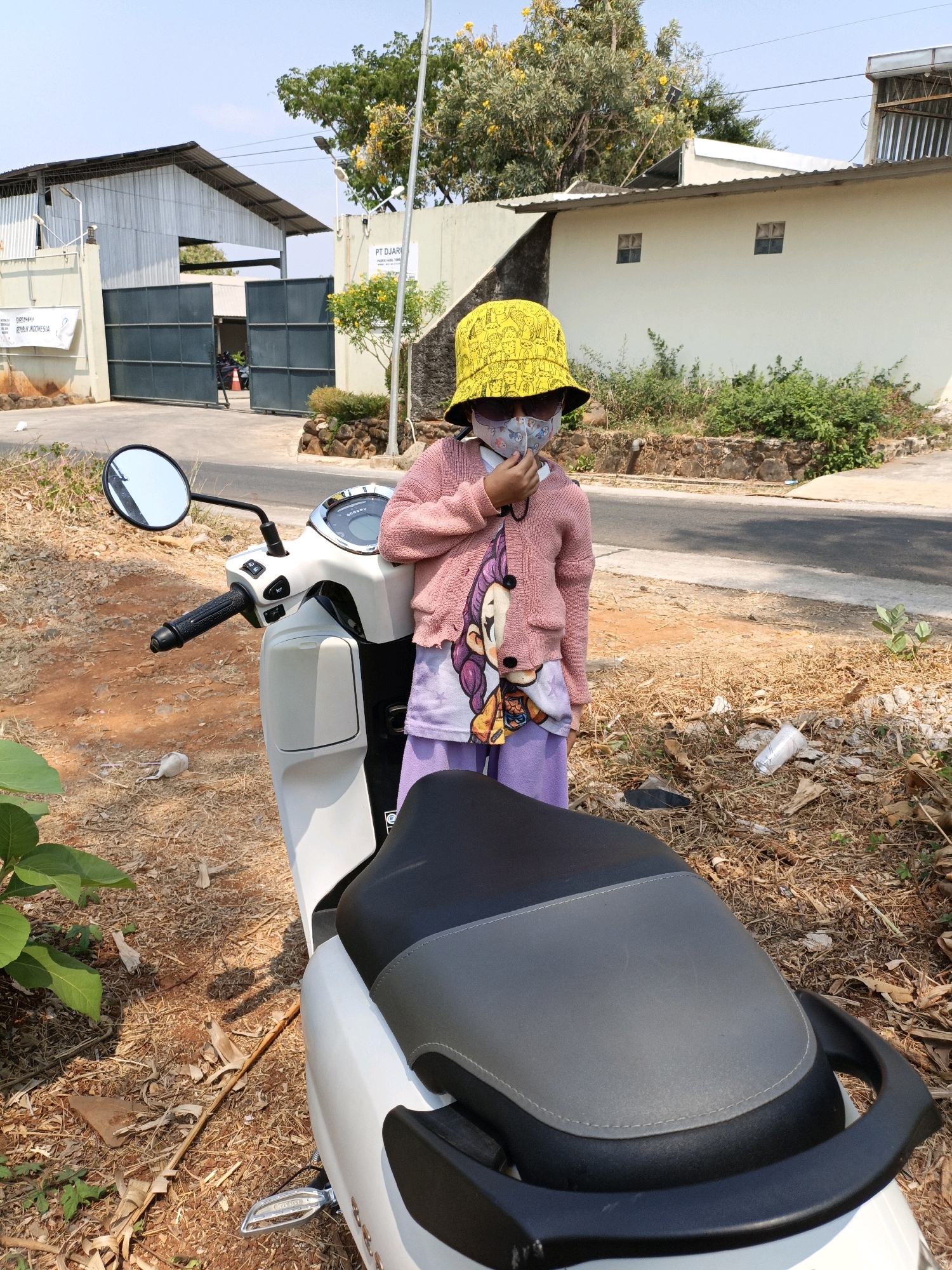 A young child wearing a yellow bucket hat, sunglasses, and a face mask stands next to a white scooter on a dirt road. The child is wearing a pink cardigan over a t-shirt and purple pants. The scooter has a black and gray seat. In the background, there is a building with a large metal gate and a sign that reads "PT DJARIM." There are trees and dry grass surrounding the area. The sky is clear and blue. The location is Kuwasen, Indonesia.