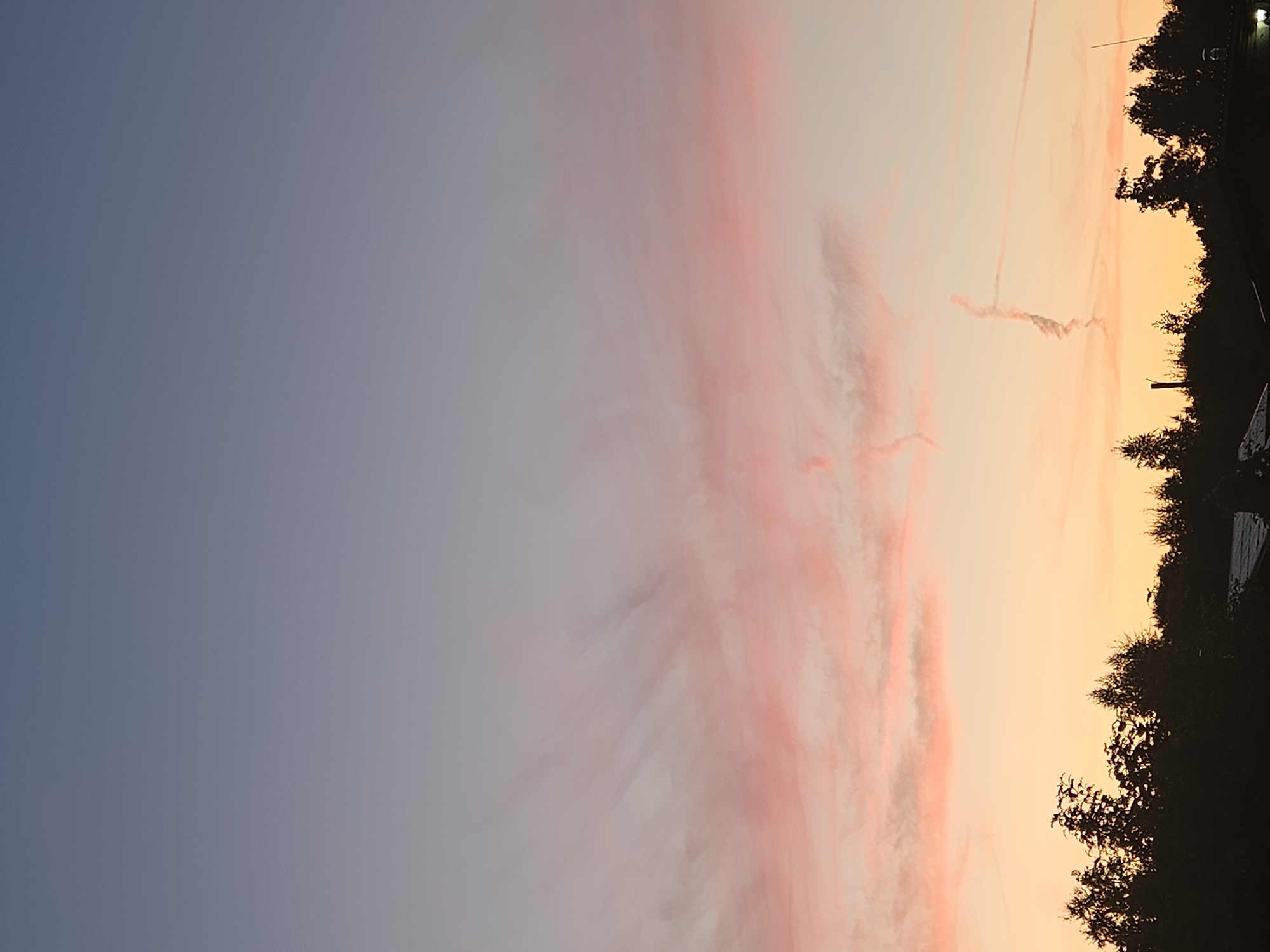 This is a full-frame, vertical, eye-level, medium shot of a sunset in the city of Kurovo, Russia. The sky is filled with wispy clouds that are tinged with pink and orange. The horizon is lined with a silhouette of trees, and a small part of a roof is visible in the bottom right corner. The overall mood is peaceful and serene.