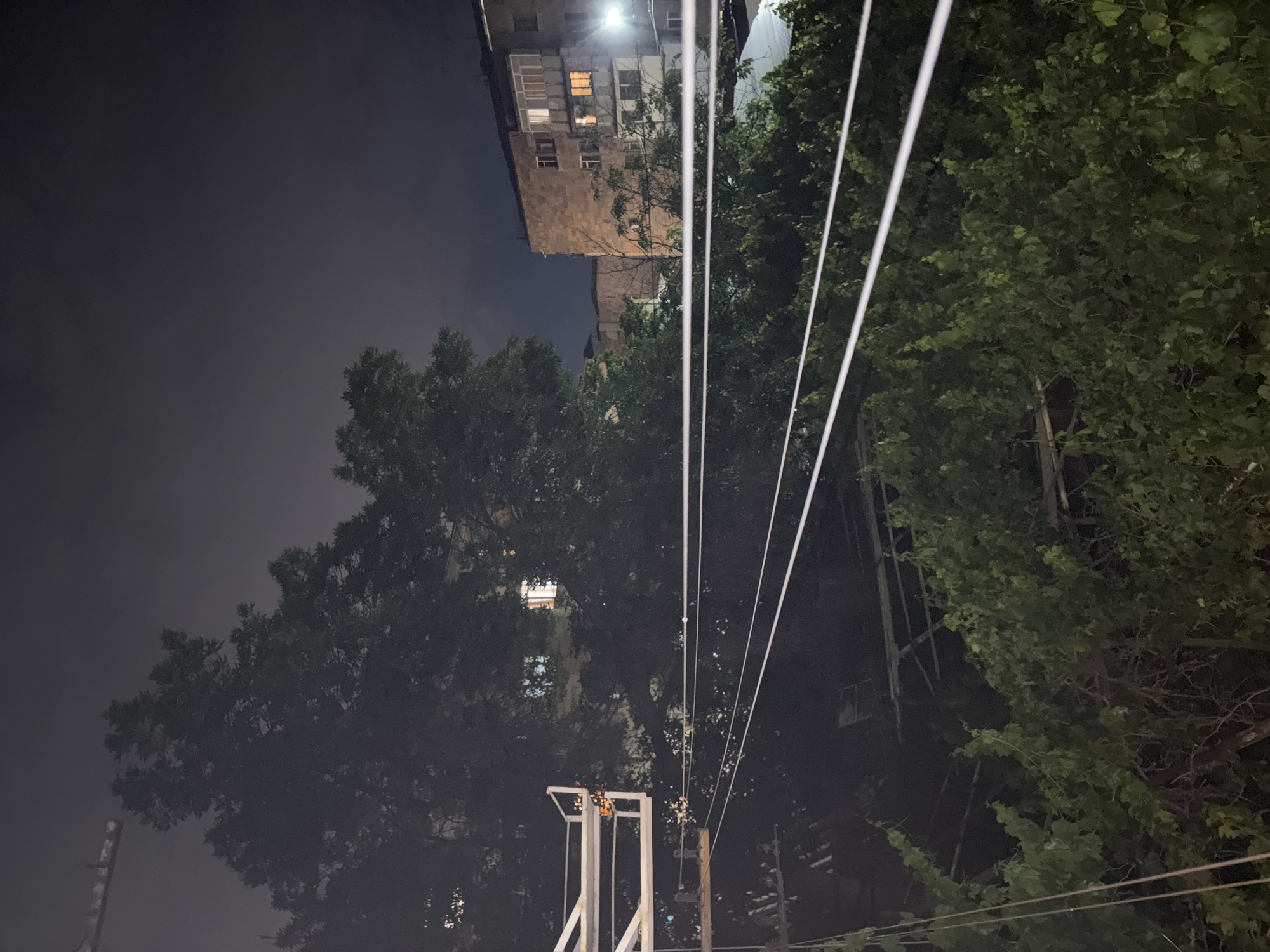 The image displays a nighttime urban scene, likely in Vagharshapat, Armenia, captured looking upwards towards the sky and surrounding elements. The sky, occupying a large portion of the frame on the left and upper parts, is dark and overcast, with a hazy, indistinct quality.
A multi-story building, appearing to be an apartment block constructed from brick or stone, is prominent in the upper right and middle sections of the image. Several windows within this building are lit with warm, artificial light, indicating occupancy. A bright streetlight shines intensely near the top of the building, illuminating parts of its facade and the nearby treetops.
Dense, green foliage from large trees fills a substantial part of the frame, particularly on the right side and across the bottom-middle. The trees partially obscure the buildings and the night sky, their leaves distinctly visible despite the low light conditions.
Multiple bright white lines, identifiable as power or communication cables, stretch diagonally across the image. These cables are supported by a white, angular metal structure positioned in the lower-middle, as well as a more traditional wooden utility pole visible to the right of the metal structure.
The scene conveys a sense of a quiet, inhabited urban environment after dark, characterized by a blend of residential architecture, natural greenery, and utility infrastructure. There are no visible people or discernible activities taking place. No visible text is present in the image.