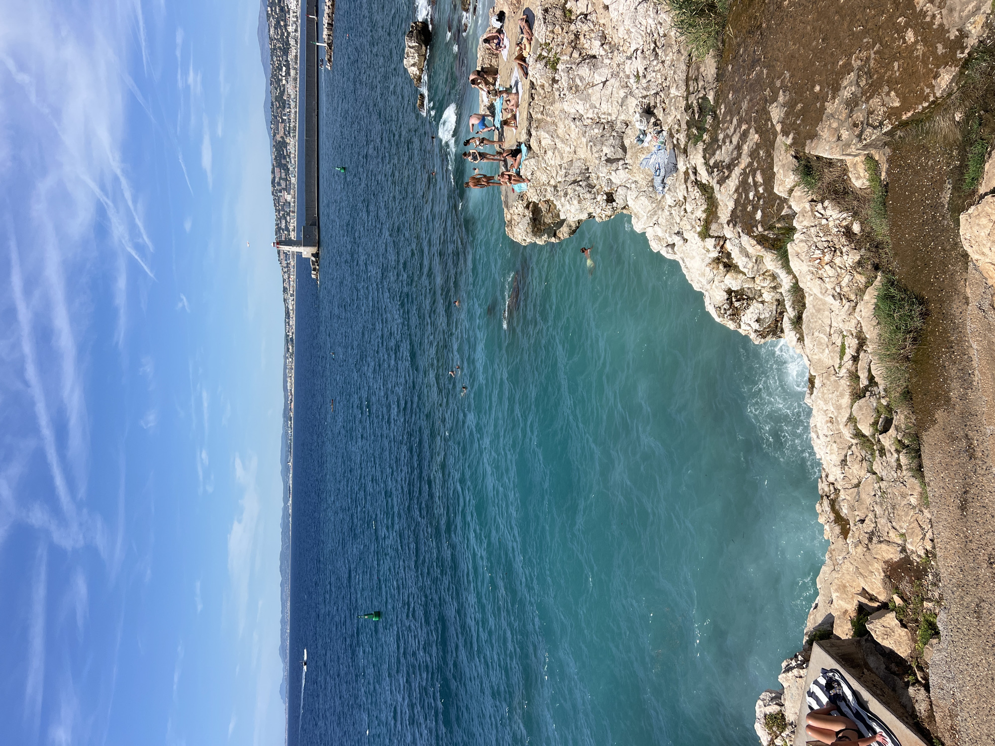 The image depicts a scenic view of the Mediterranean Sea in Nice, France. In the foreground, a rocky coastline is visible with several people sunbathing and relaxing on the rocks. The sea is a vibrant turquoise color, with waves gently crashing against the shore. In the background, a long pier extends into the water, with a lighthouse at its end. The city of Nice is visible along the coastline, with buildings stretching as far as the eye can see. The sky is a clear blue with a few wispy clouds. The overall impression is one of a beautiful and tranquil day at the beach.