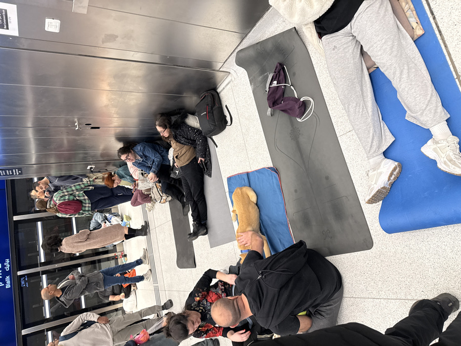 The image depicts an indoor scene within the Bialik Light Rail Station in Ramat Gan, Israel, identified by a visible "Bialik" sign on a glass partition in the background. Approximately 15-20 individuals, primarily adults, are present in various states of rest. The floor is composed of light-colored tiles.
In the foreground and midground, multiple individuals are seated or lying on dark gray and blue floor mats. A man in a black t-shirt is seated on the floor, petting a light brown dog that is lying on a blue mat. A woman in a black and red floral top is seated nearby. To the right, a woman is lying prone on a gray mat, her head resting on a pillow, adjacent to a black backpack and a reflective stainless steel wall. Another woman sits upright against this wall, holding a small object.
In the background, several individuals are standing and conversing near a glass partition, behind which a vacant platform or track area is visible. The stainless steel wall on the right features two doors and a small white wall-mounted device. Various personal belongings, including backpacks, bags, and articles of clothing, are scattered among the individuals. The overall scene suggests a temporary communal resting or sheltering area within the transit station.